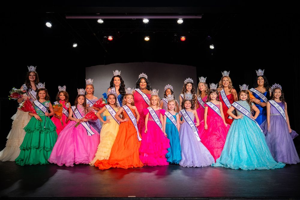 Large group of pageant contestants in colorful formal dresses pose together on dark stage.