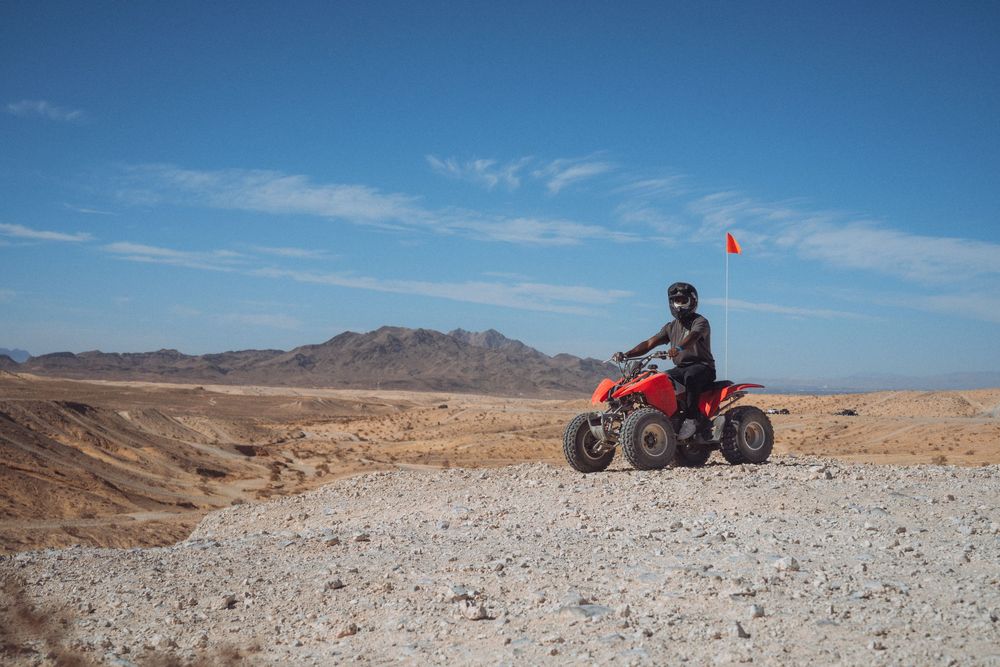 Off-road vehicle traverses rocky desert terrain under bright blue skies with mountains in the distance.