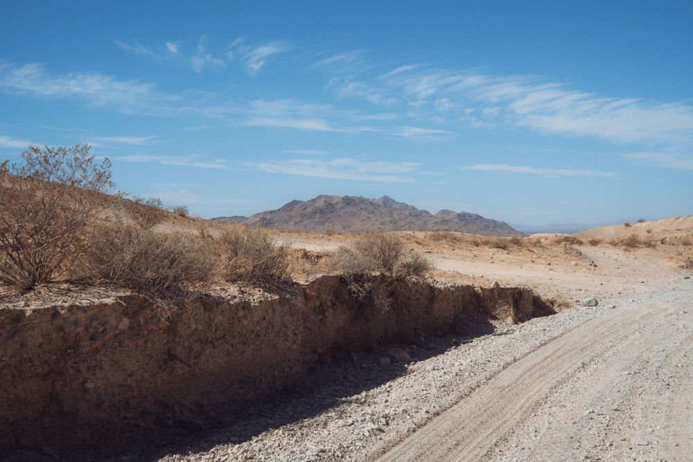 Winding dirt road cuts through desert terrain with mountains and brush visible.