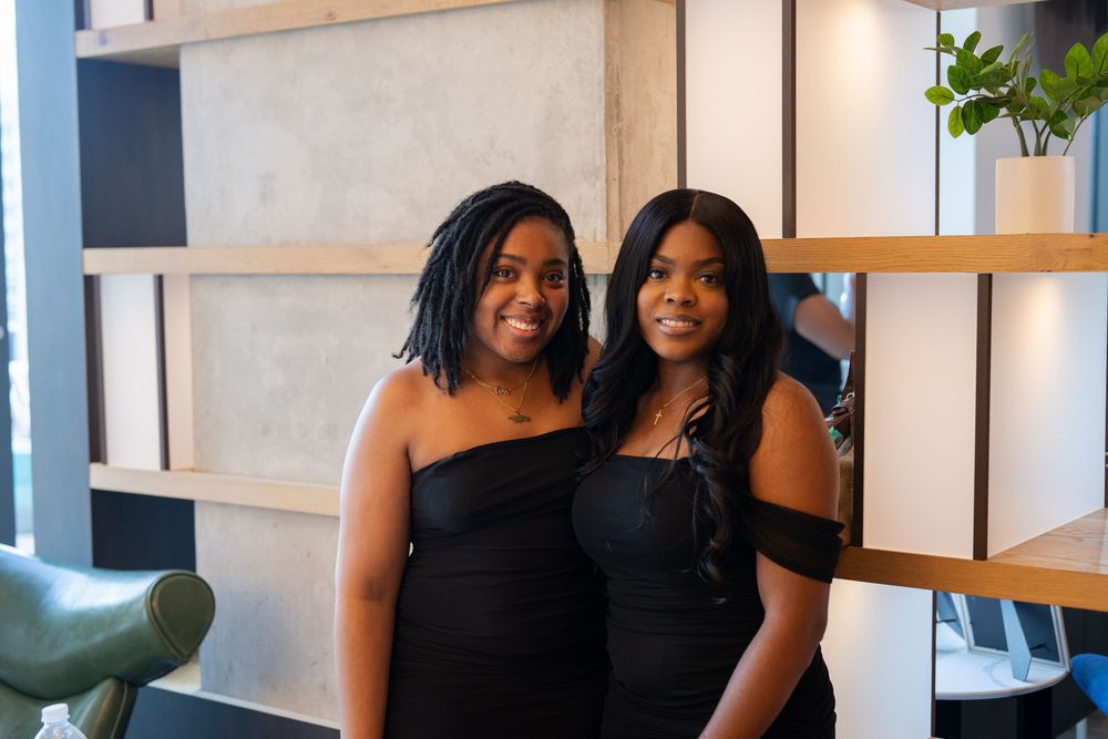 Two women in black dresses smile warmly at a social gathering.