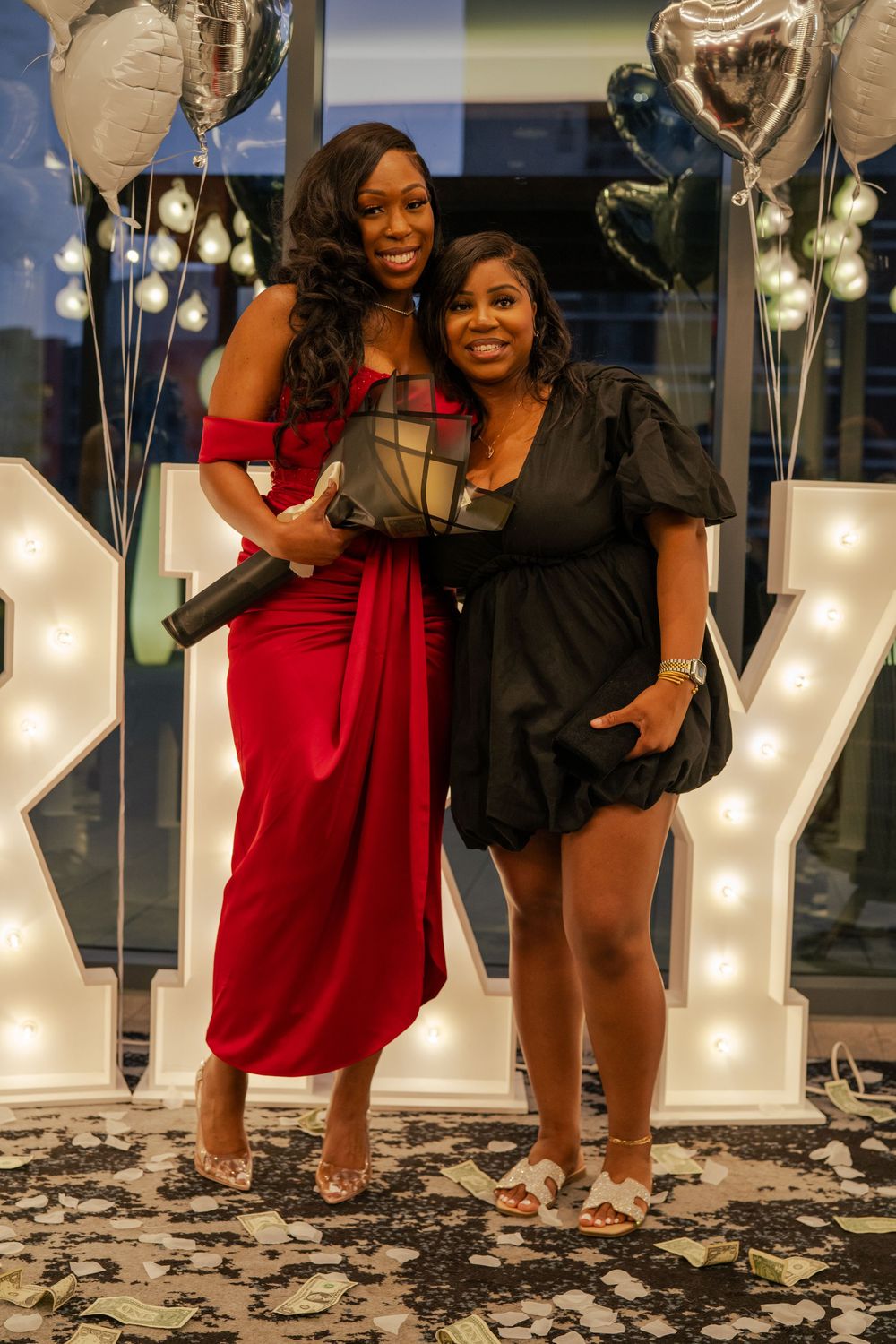 A couple poses with a decorative money bouquet at an elegant event with string lights in the background.