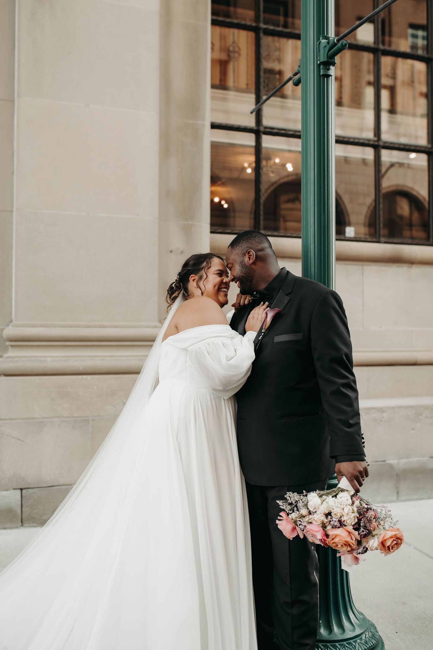 A bride and groom share a romantic moment near a green lamppost outside a grand building.