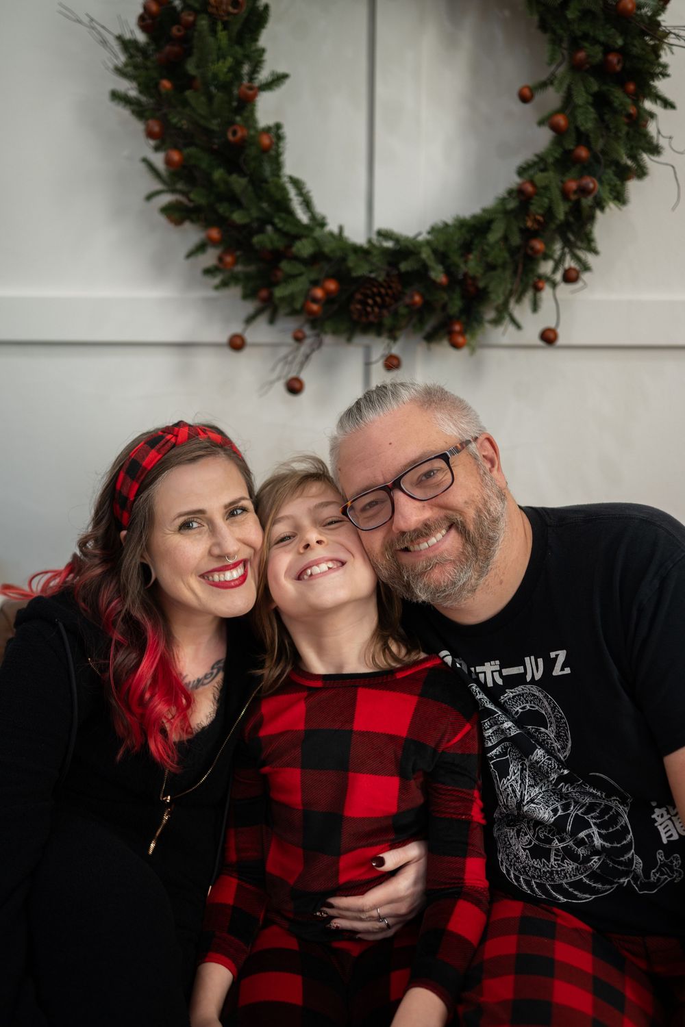 A family wearing coordinating red buffalo plaid and black outfits poses for holiday photos with a wreath backdrop.