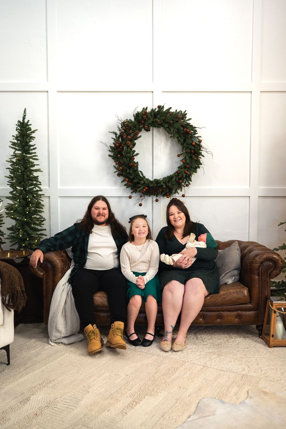 Three people sit together on floor against white wall with Christmas decorations and brown leather couch visible.