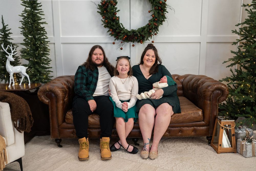 Group sitting on brown leather couch with holiday decorations and evergreen wreath hanging on white wall behind them.