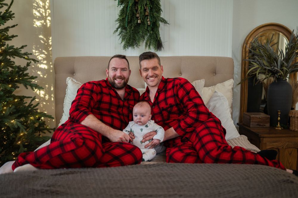 Family wearing matching red and black buffalo plaid pajamas poses on bed with Christmas tree in background.