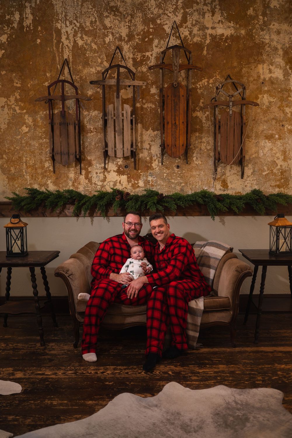 Family wearing matching red and black buffalo plaid pajamas sits on rustic wooden bench with holiday greenery decor.