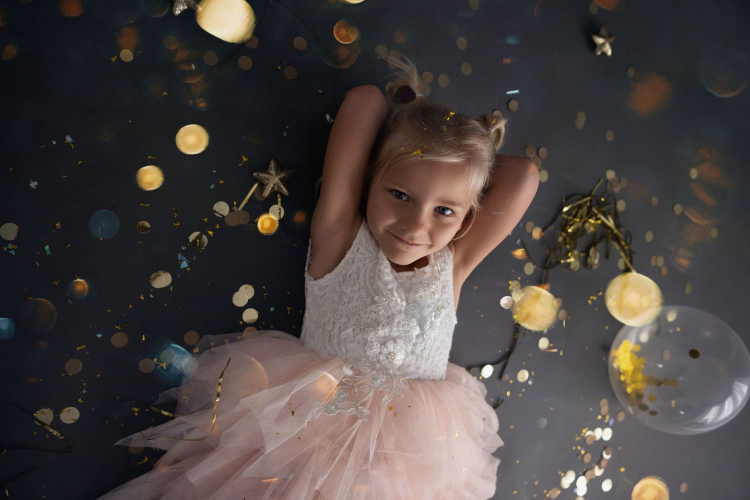Young dancer in pink tulle dress twirls beneath falling gold glitter and sparkles against dark background.