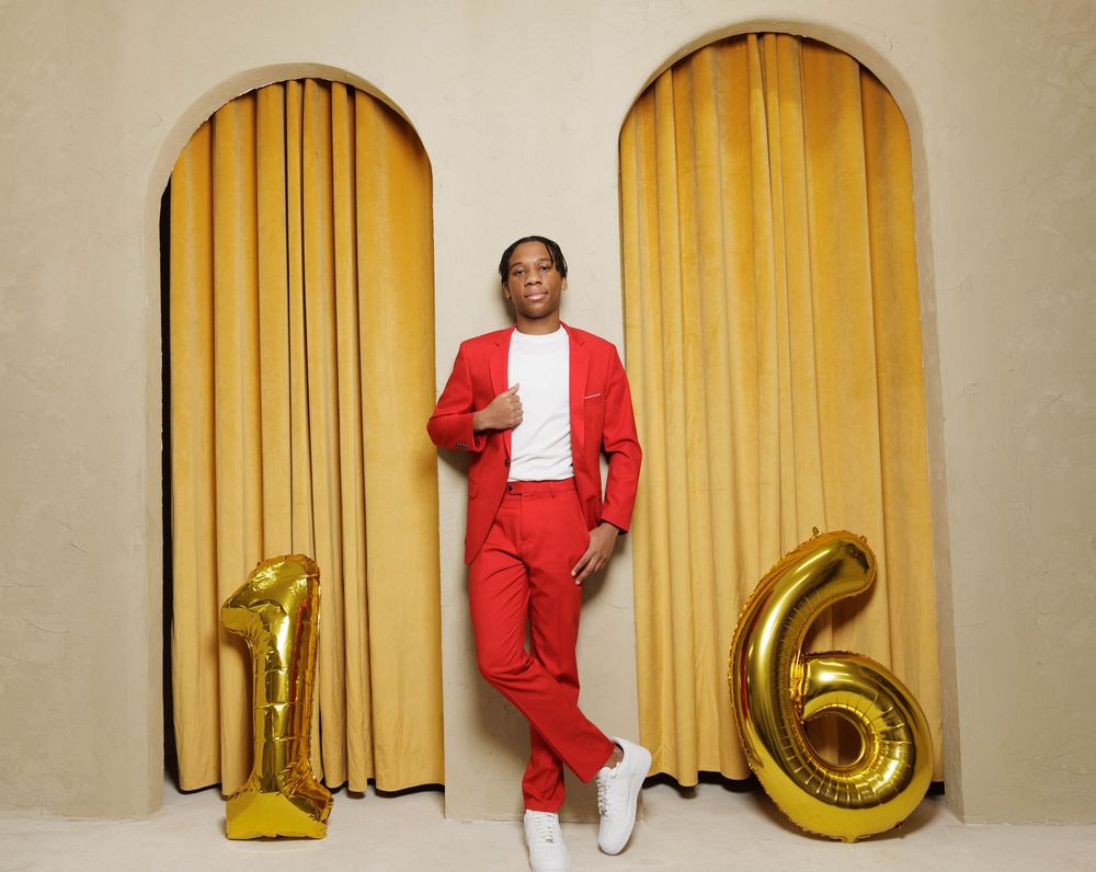 A person in a bright red suit poses in front of gold curtains in a stylish fashion photo series.