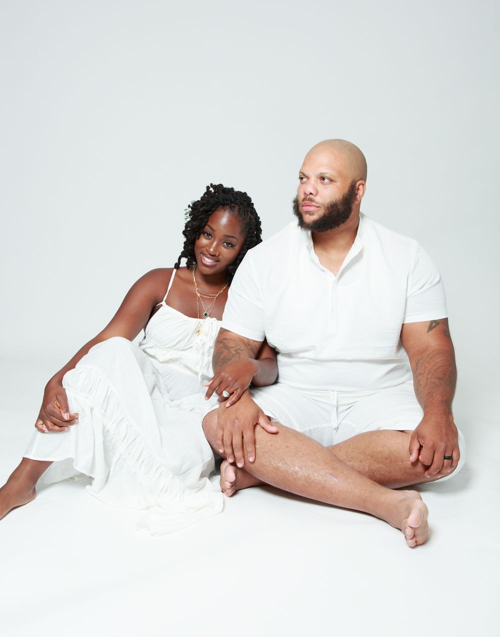 Couple in white outfits poses intimately on white background for minimalist studio portraits.