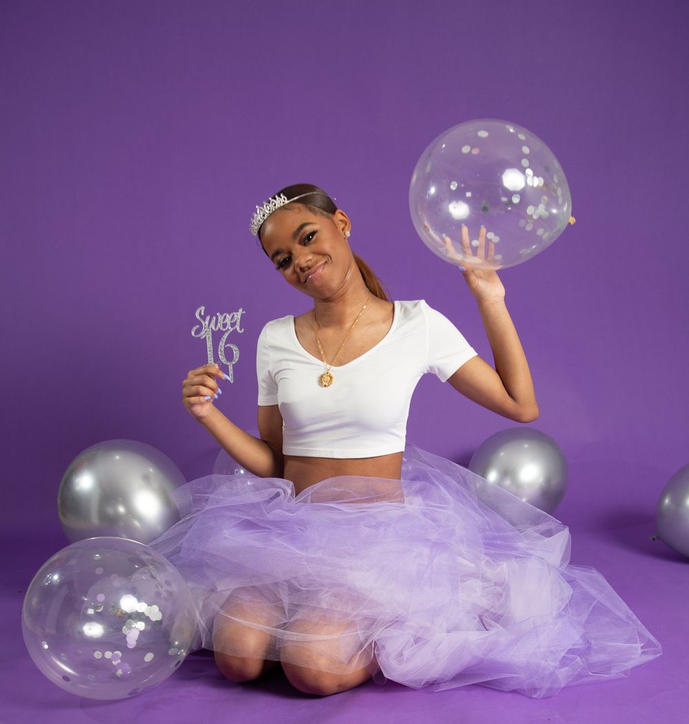 A person in a white crop top and lavender tulle skirt poses with silver and clear balloons against a purple backdrop.