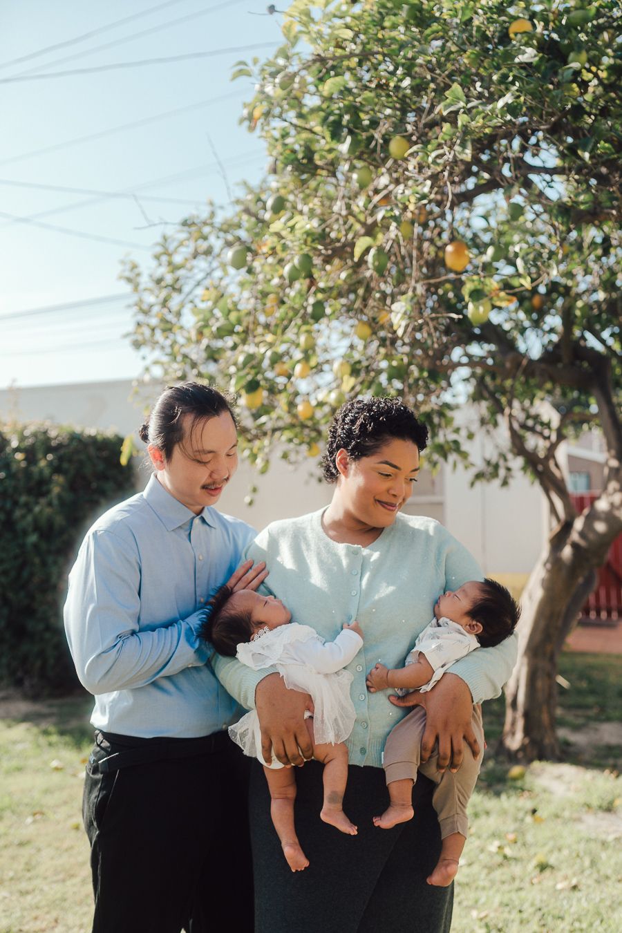 A couple in matching white and blue outfits sharing tender moments with their baby outdoors near a citrus tree.