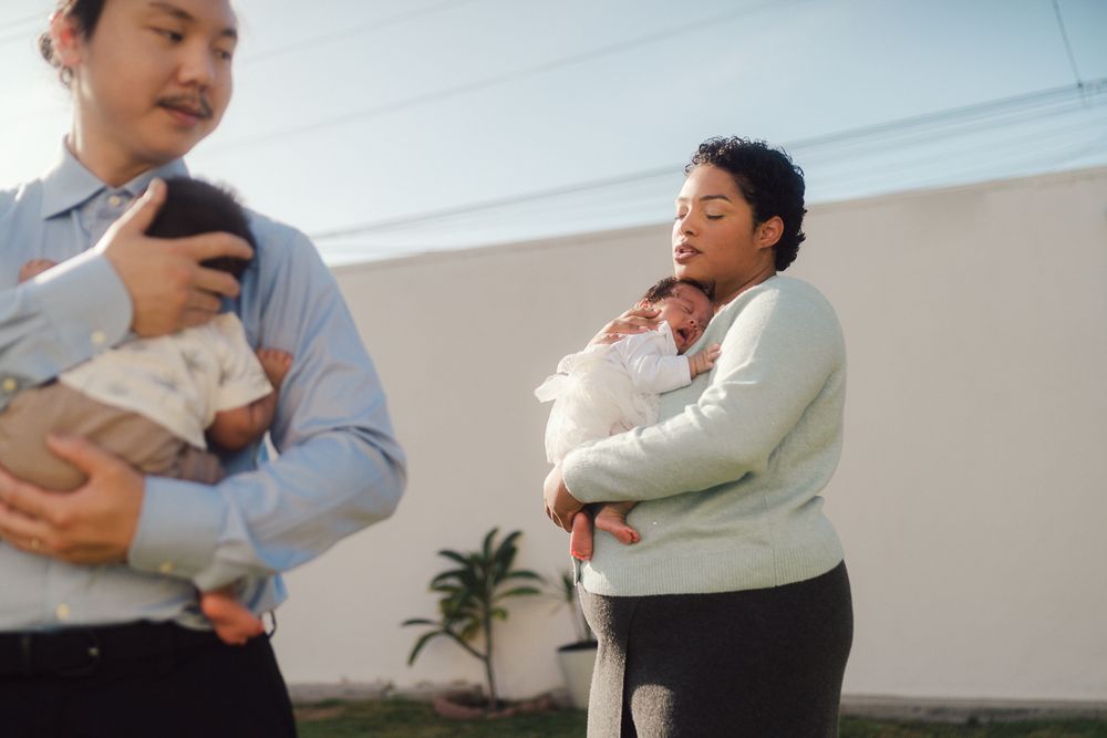 A couple in coordinated outfits holding and interacting with their infant baby against a light-colored wall.