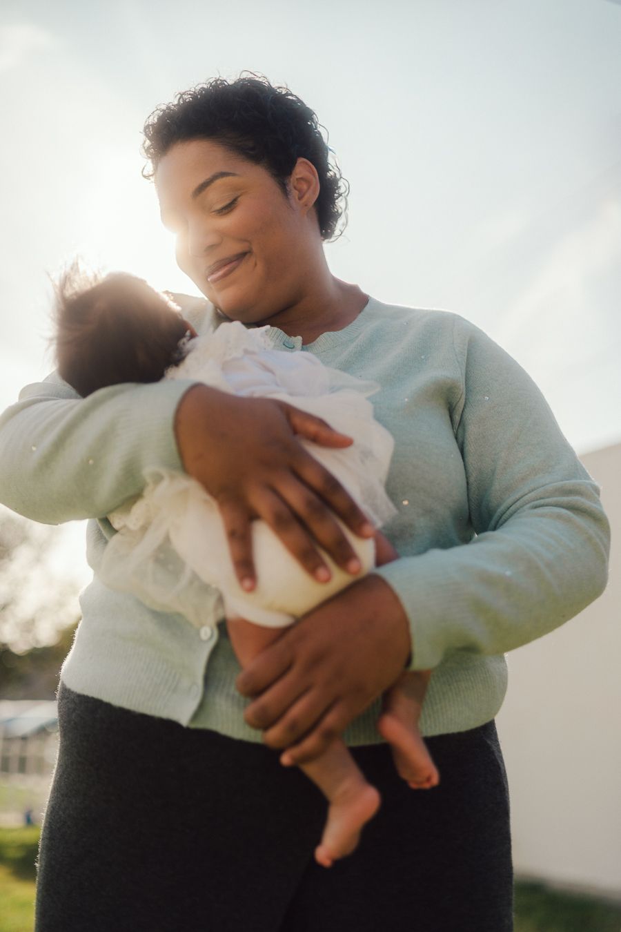 A parent in a white sweater tenderly holds and gazes at their infant against a bright outdoor backdrop.