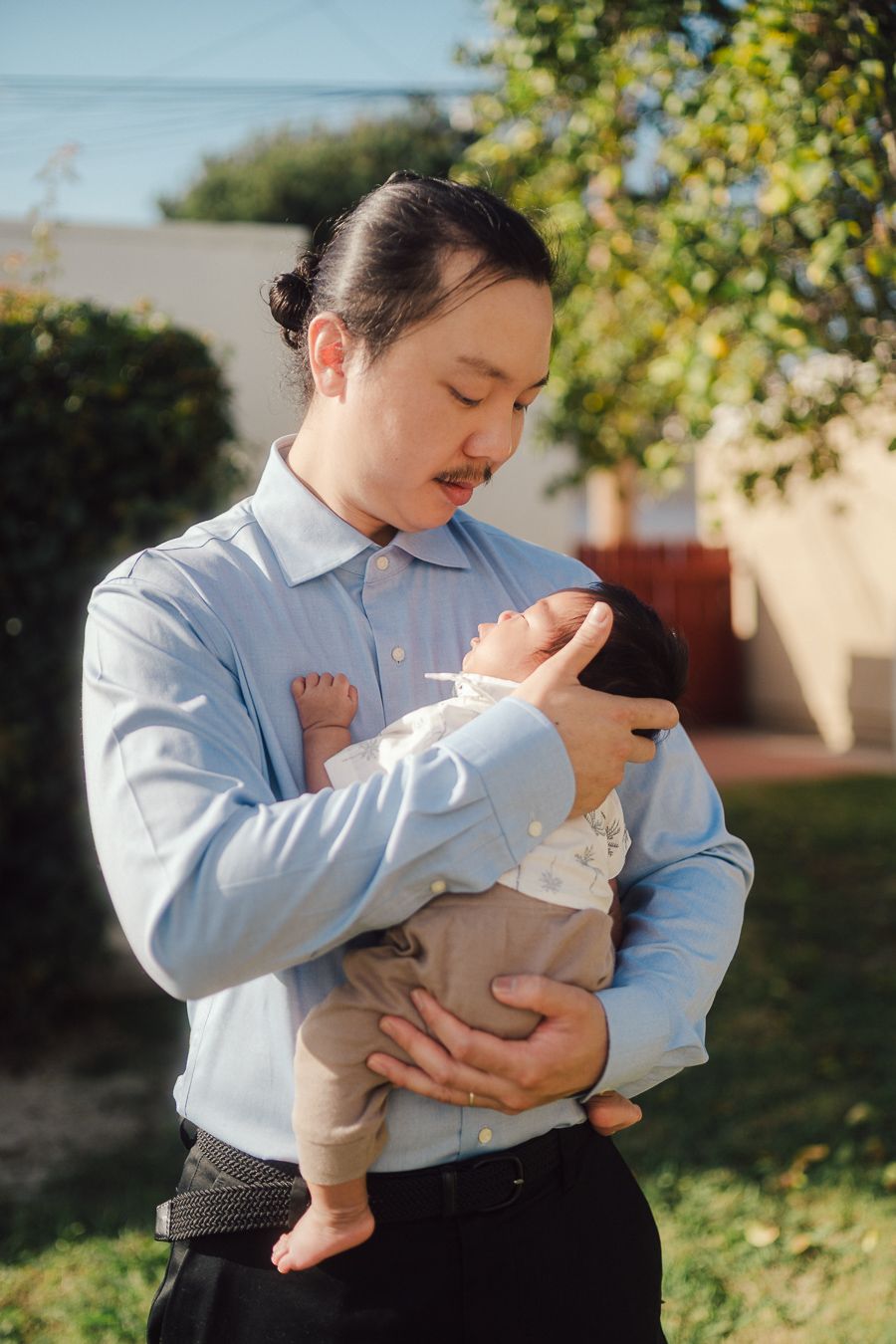 A tender moment between parent and baby in a light blue shirt against a sunny outdoor backdrop with trees.