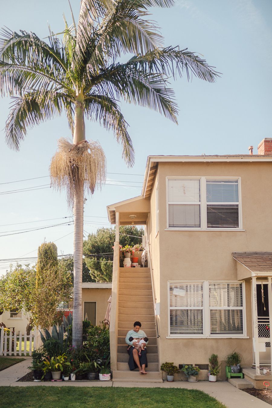 A tall palm tree stands beside a beige two-story house with someone sitting on the outdoor staircase.