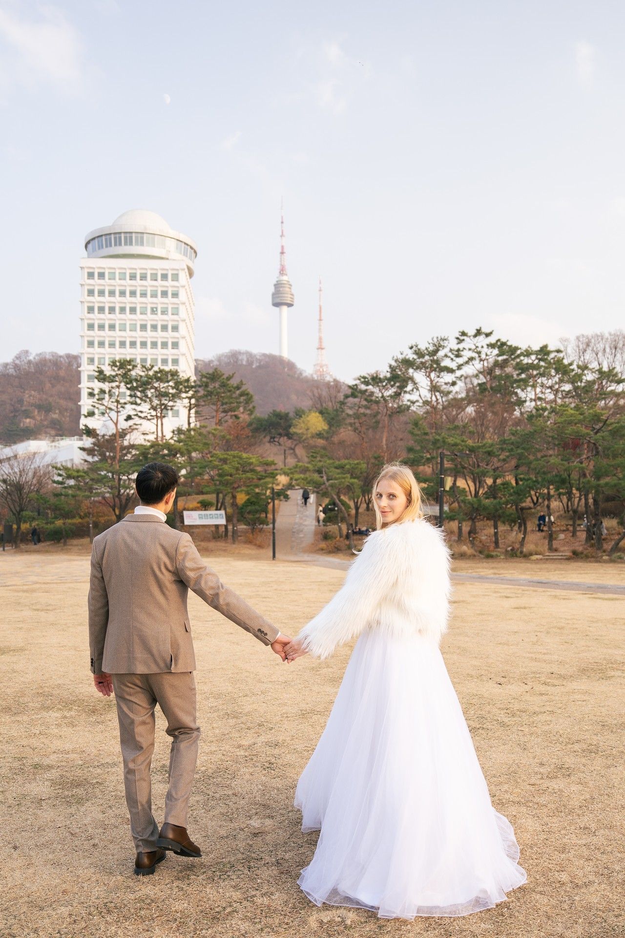 A couple in wedding attire hold hands in a park with N Seoul Tower and buildings visible in the background.