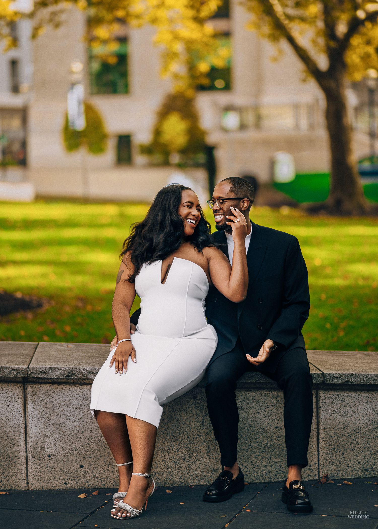 A couple in formal attire sits together on a stone ledge in a park during autumn, sharing intimate moments.