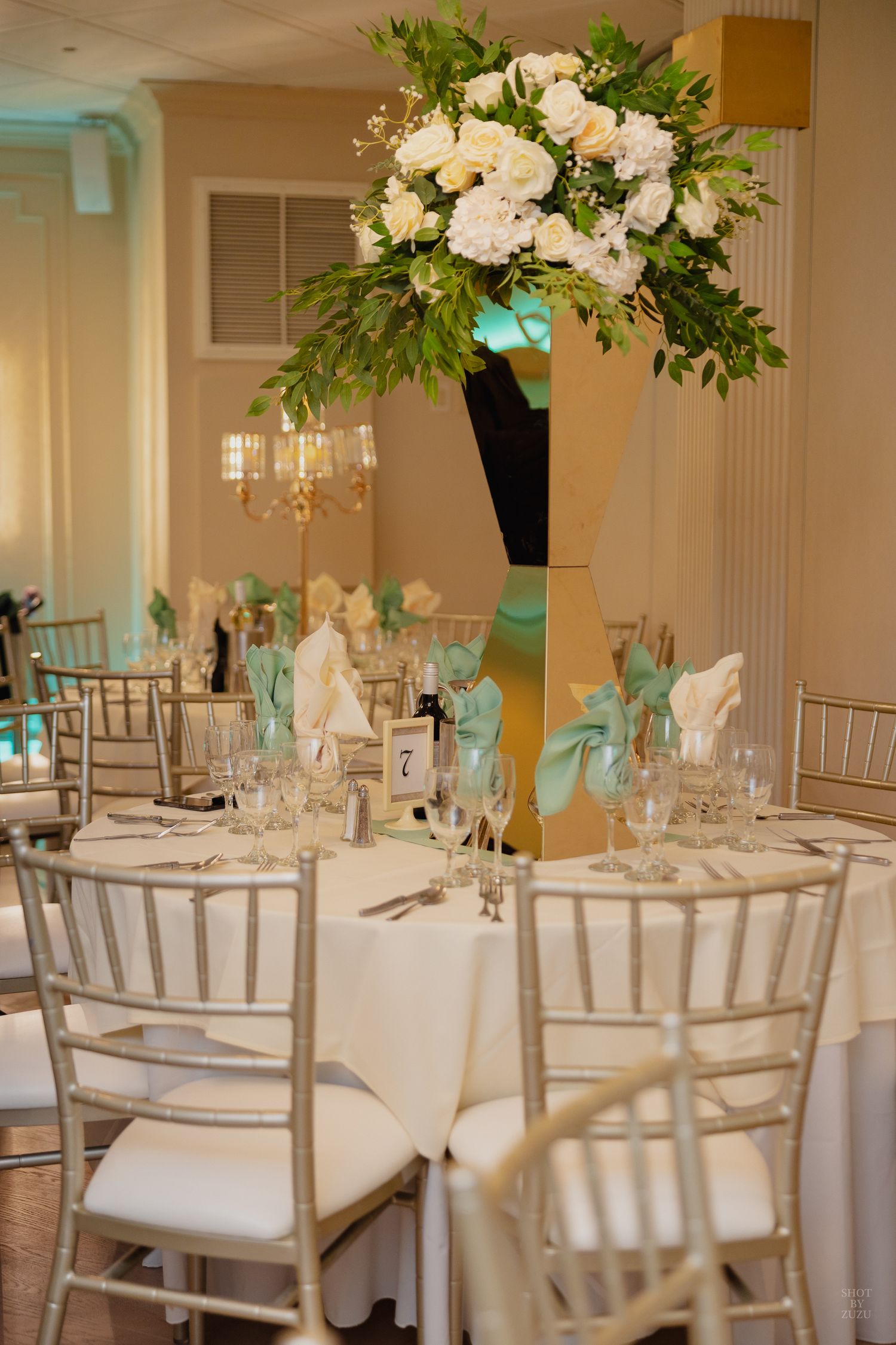 Elegant wedding reception venue with white chiavari chairs, round tables, and tall floral centerpieces in white and green.