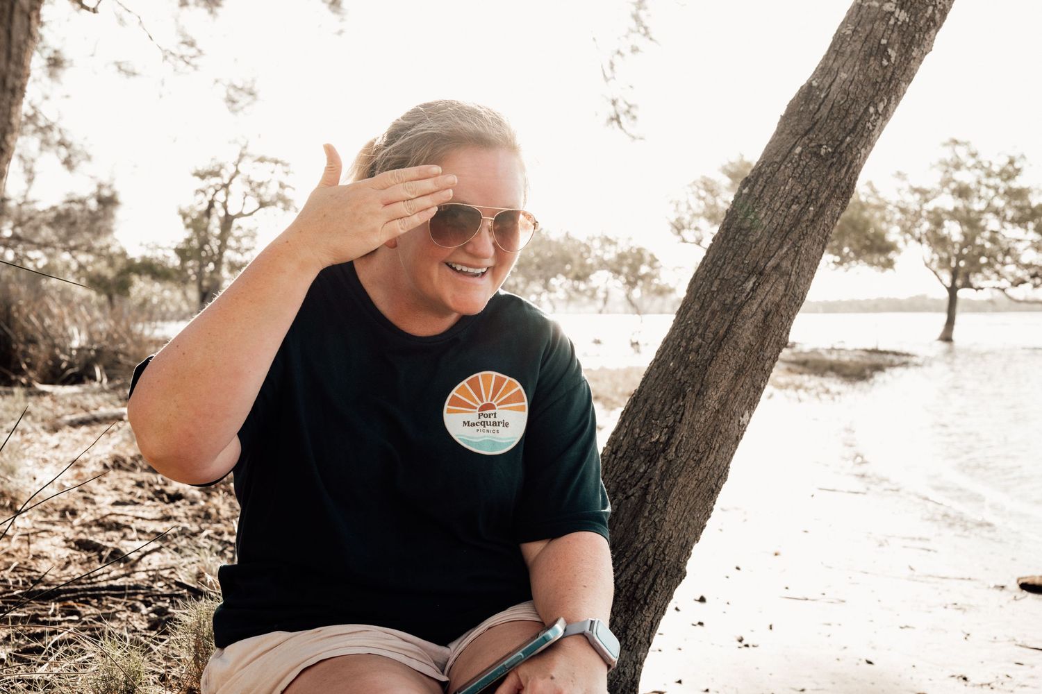 A person in a black t-shirt sits outdoors smiling while enjoying a sunny day in nature.