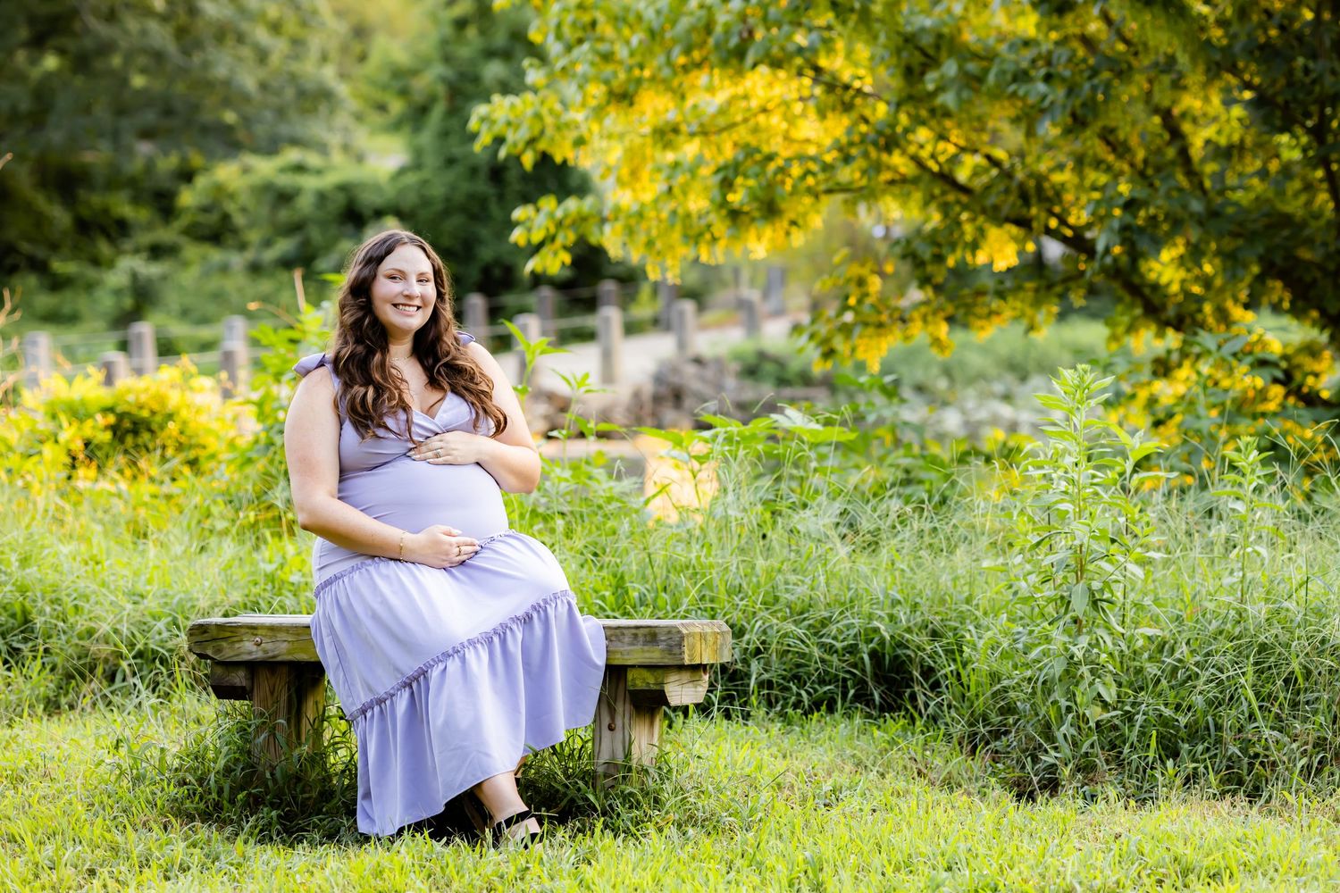 A pregnant woman in a flowing lavender dress sits on a wooden bench surrounded by blooming yellow flowers in a garden setting.