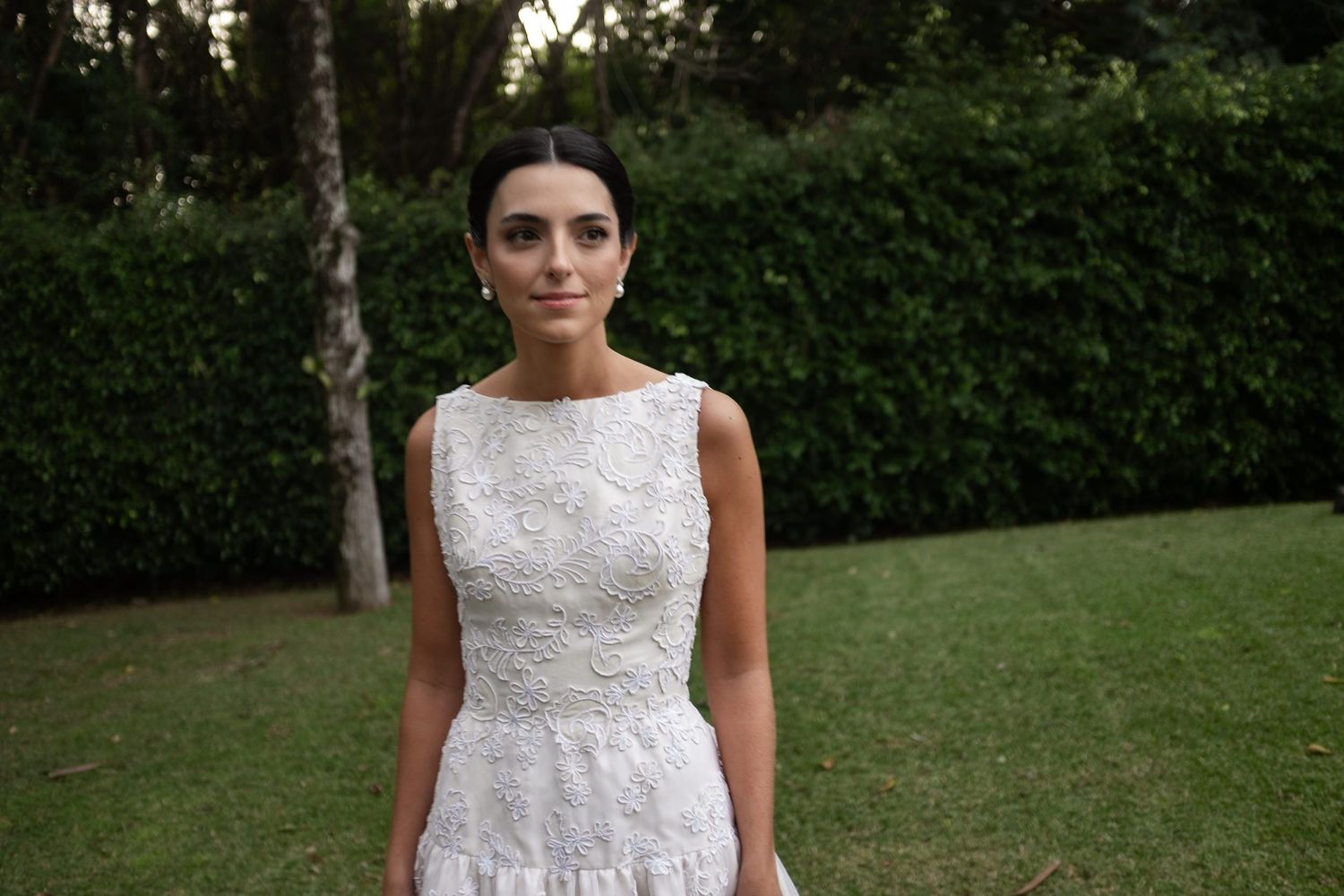A bride in a white lace sleeveless dress poses in a garden setting during golden hour.