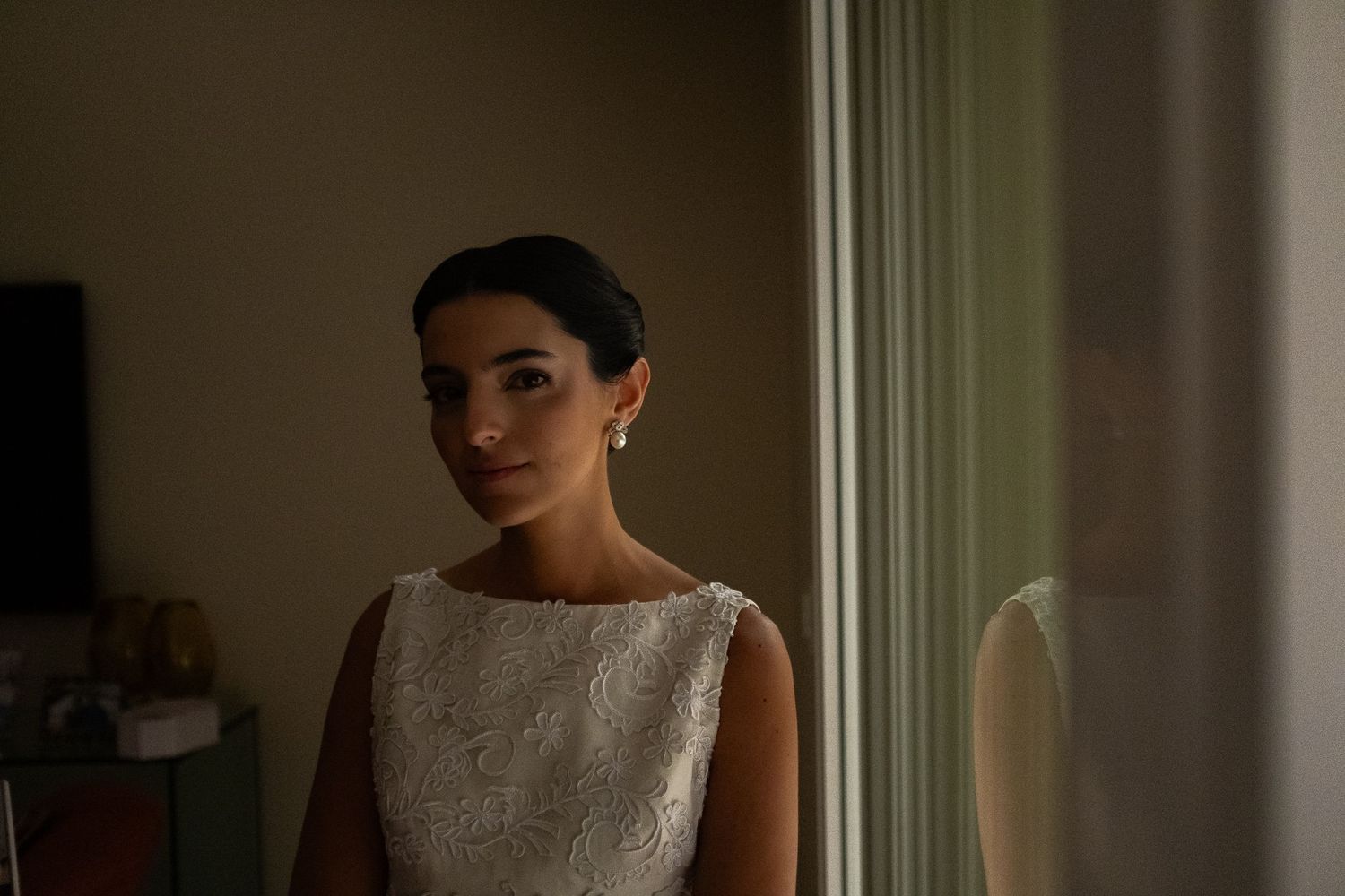 A bride in a white lace wedding dress stands by a window in soft natural light.