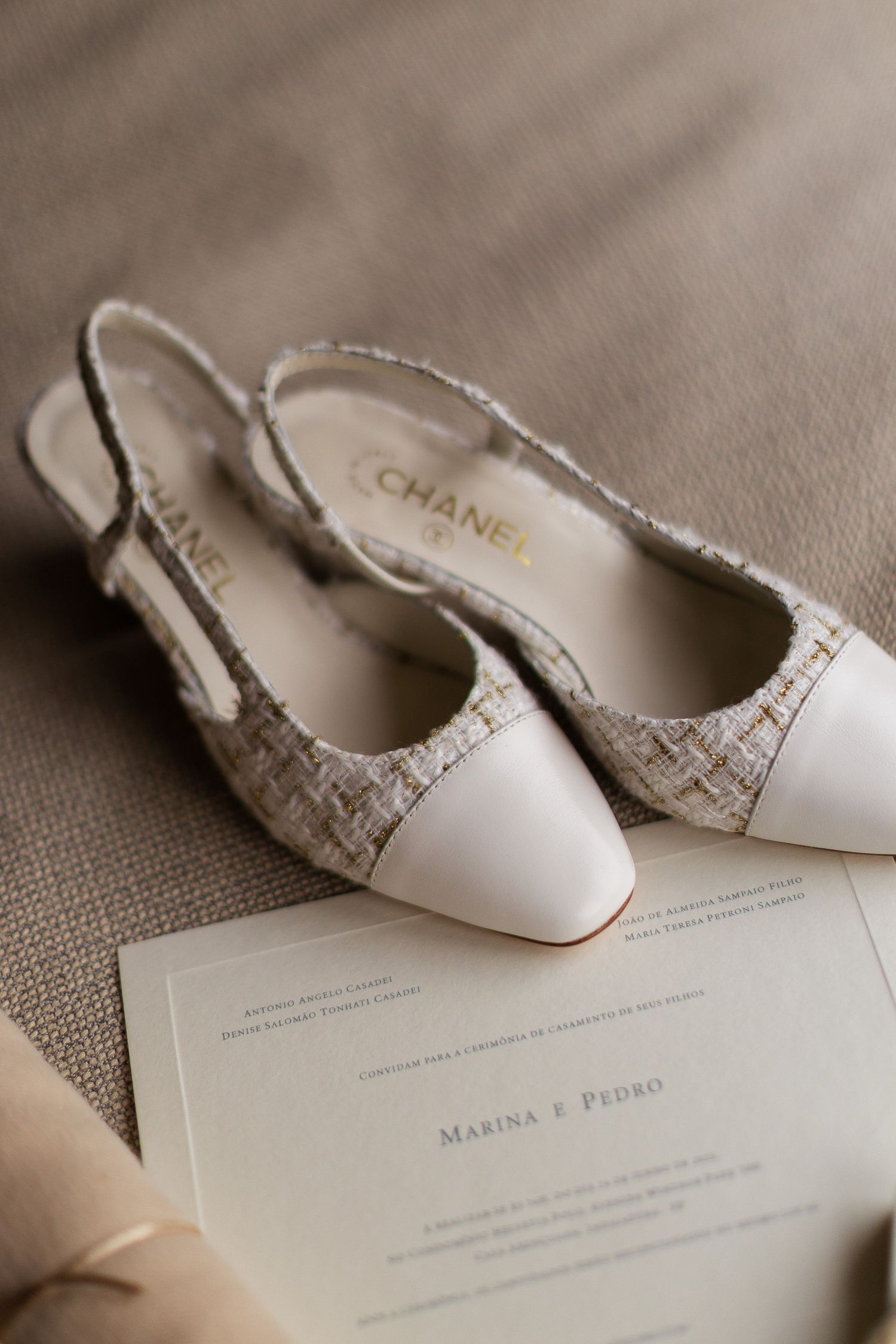 A pair of elegant Chanel slingback heels in beige with tweed detailing sits atop a wedding invitation.