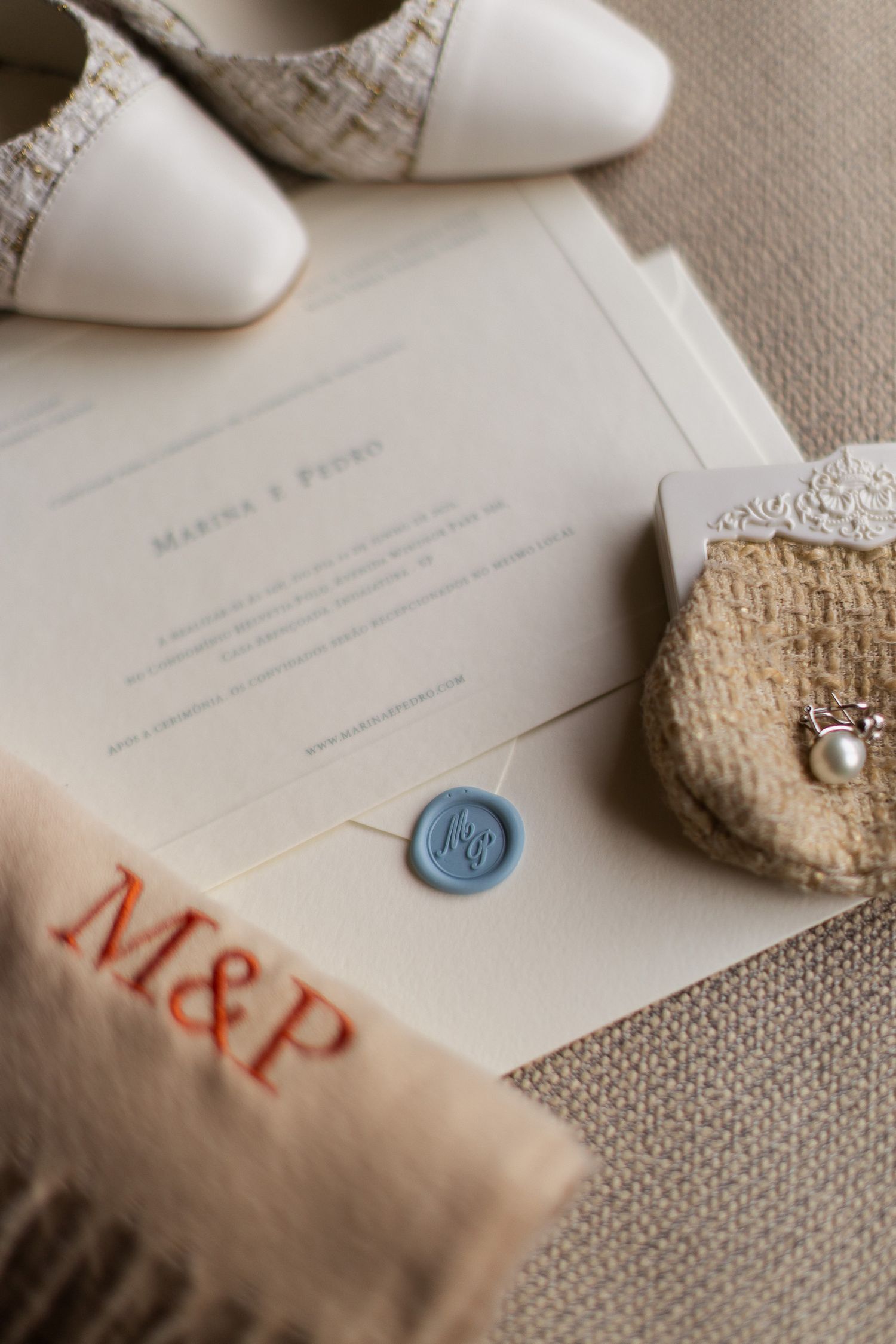 Wedding invitation with blue wax seal, white shoes, and monogrammed M&P details on textured fabric.