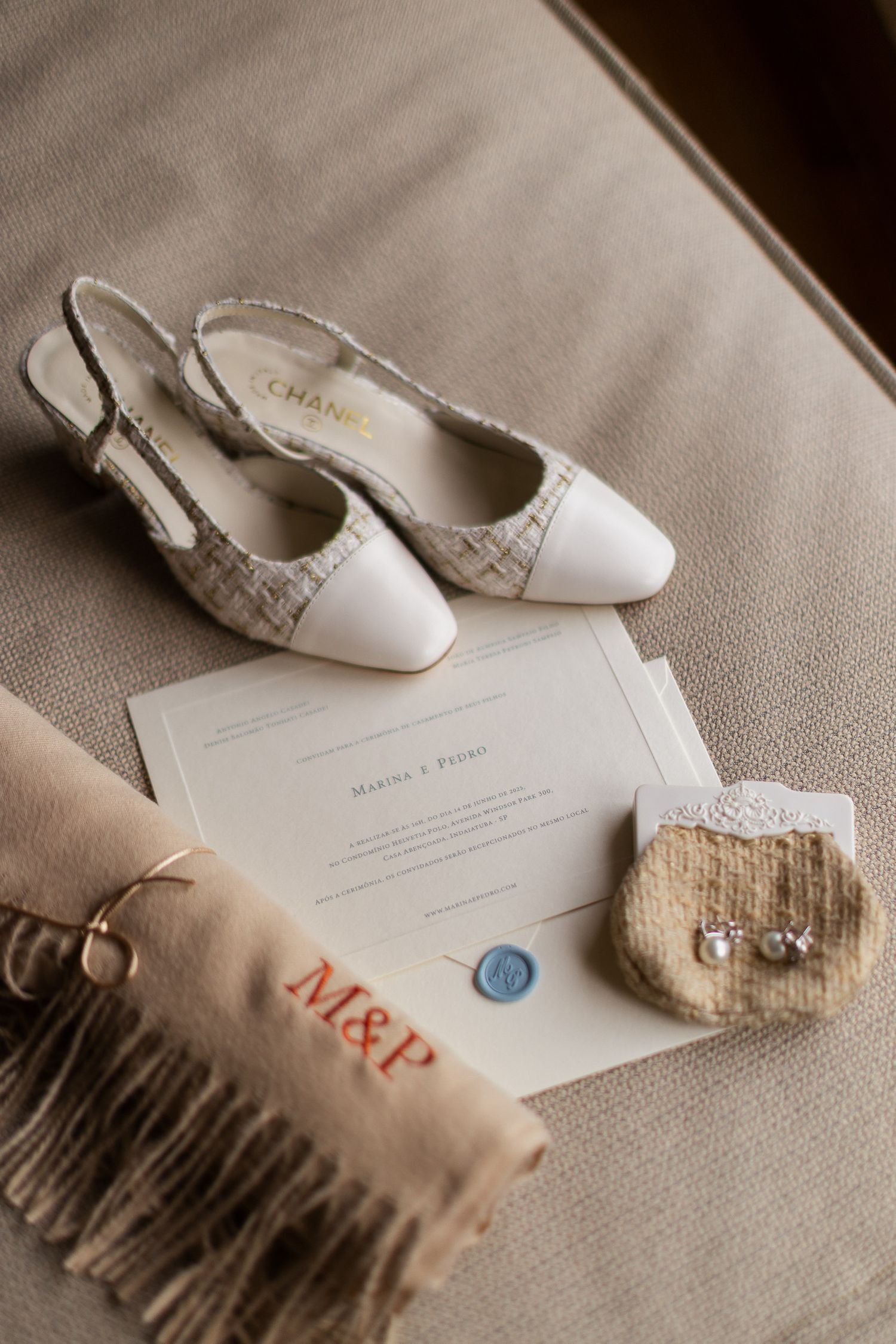 Elegant white bridal shoes and wedding invitation with blue seal displayed on beige fabric with M&P monogram.