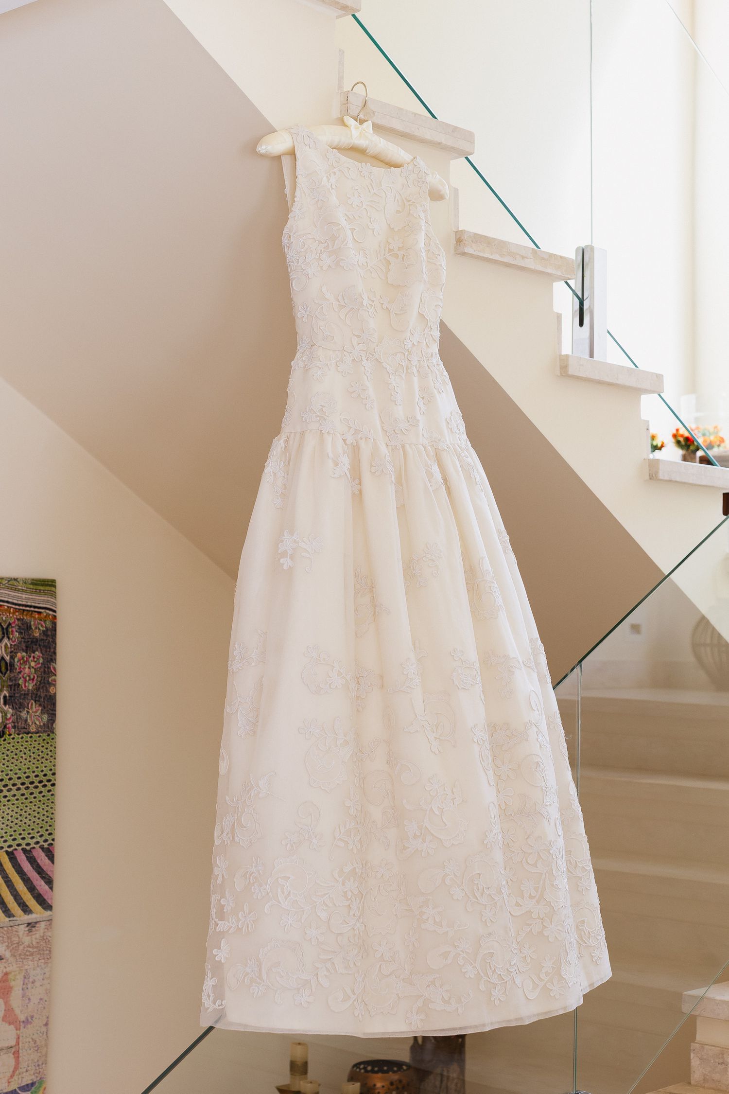White lace christening dress hanging on display against a beige wall with stairs in the background.
