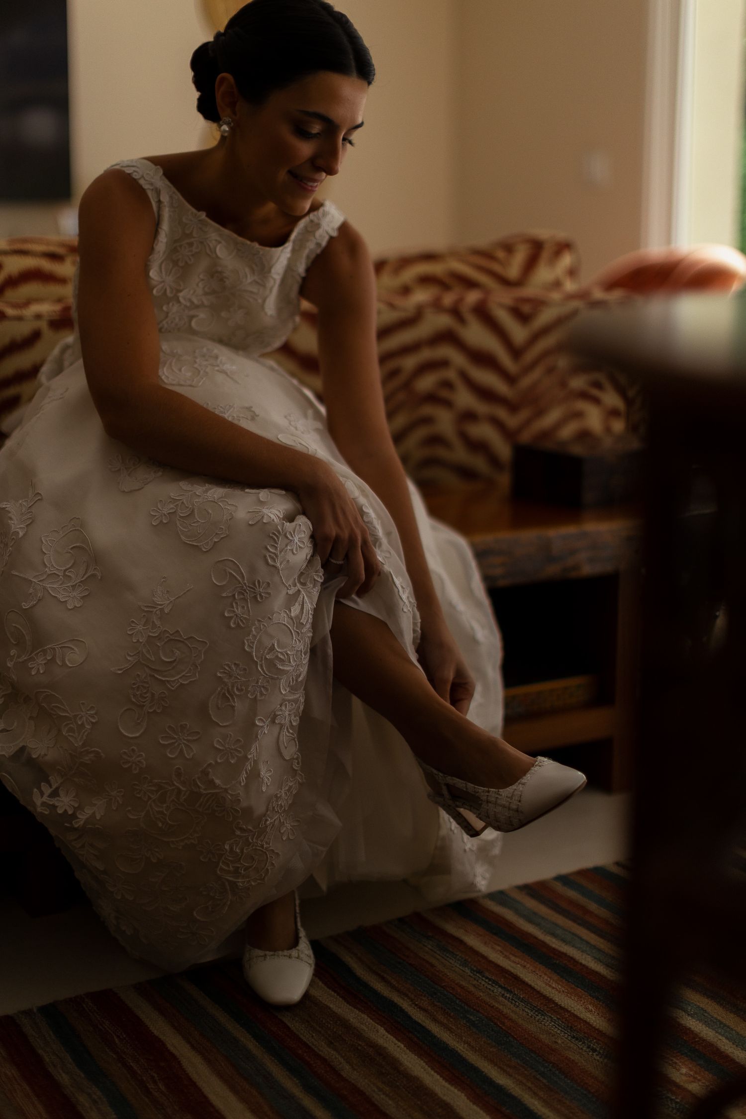 A person in a white lace wedding dress puts on white shoes while sitting on a striped couch.