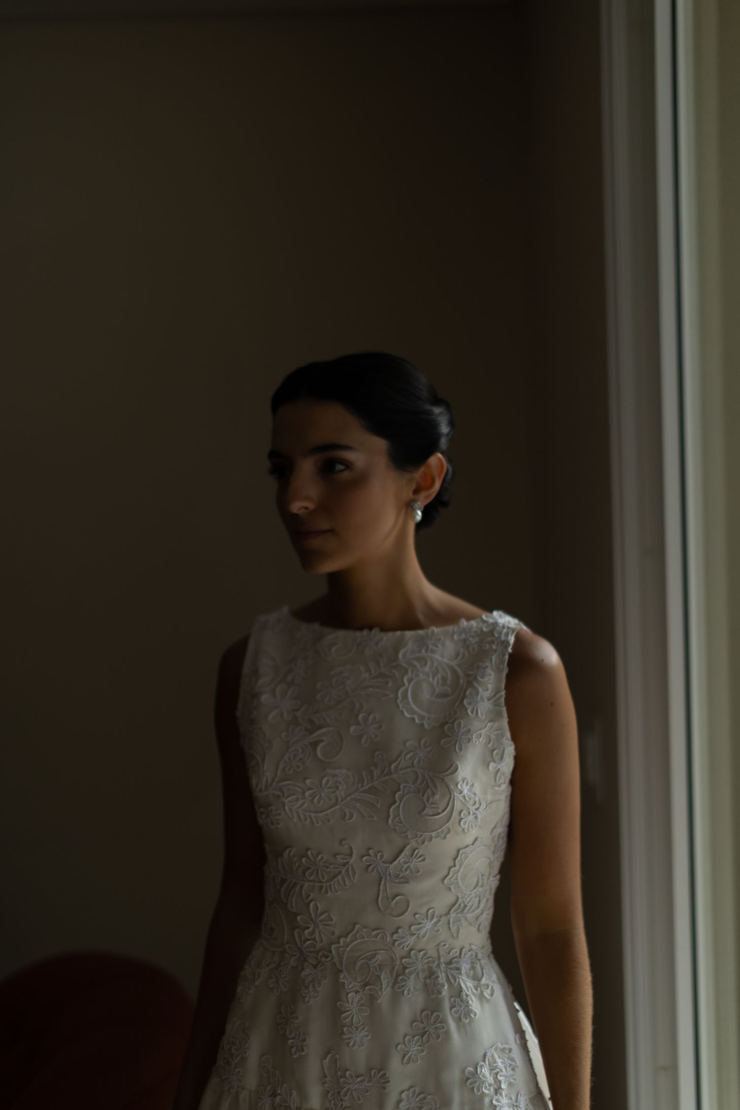 A person in a white lace sleeveless dress stands by a window in dramatic lighting.