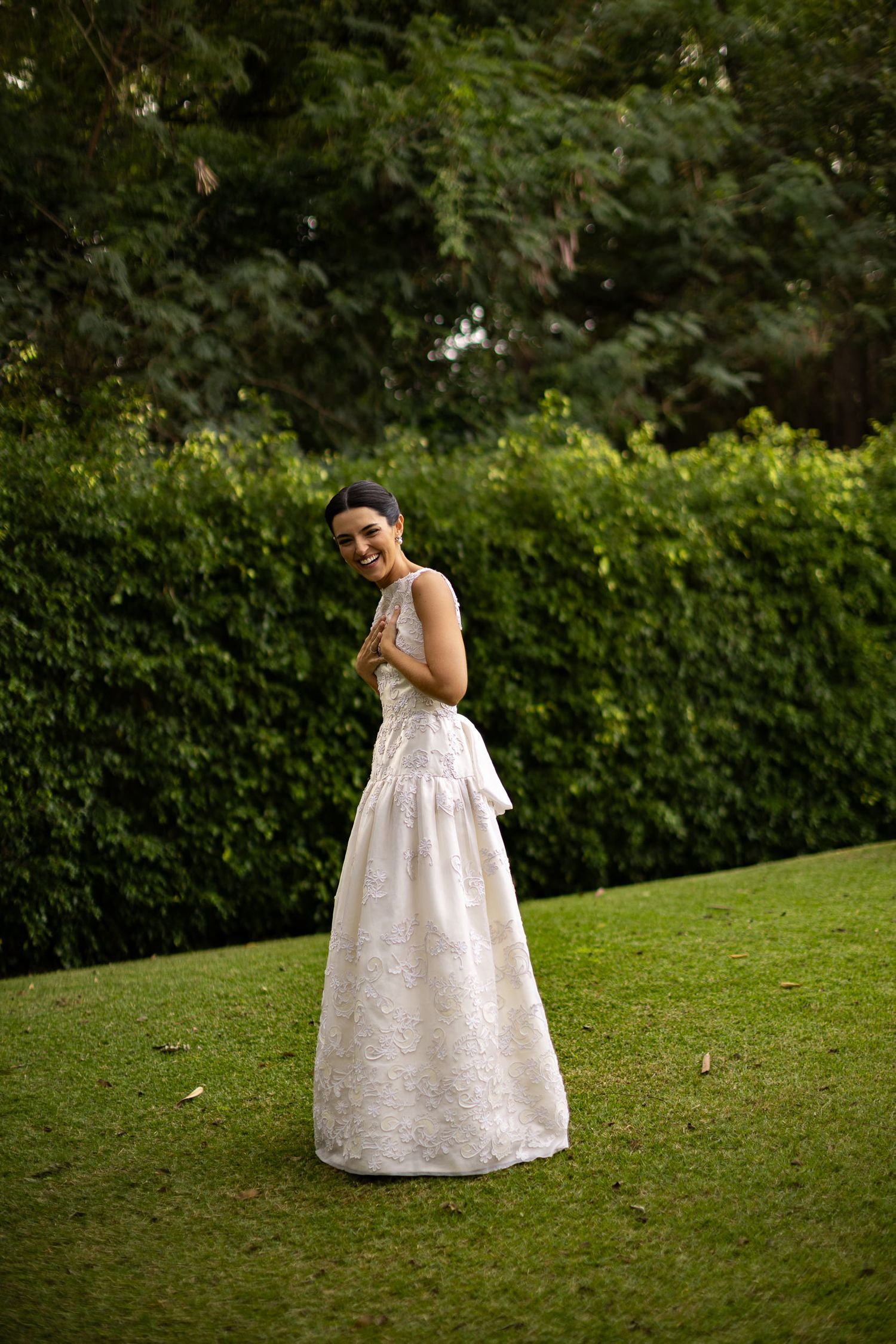 A person in an elegant white lace wedding gown poses on a manicured lawn with green hedges in the background.