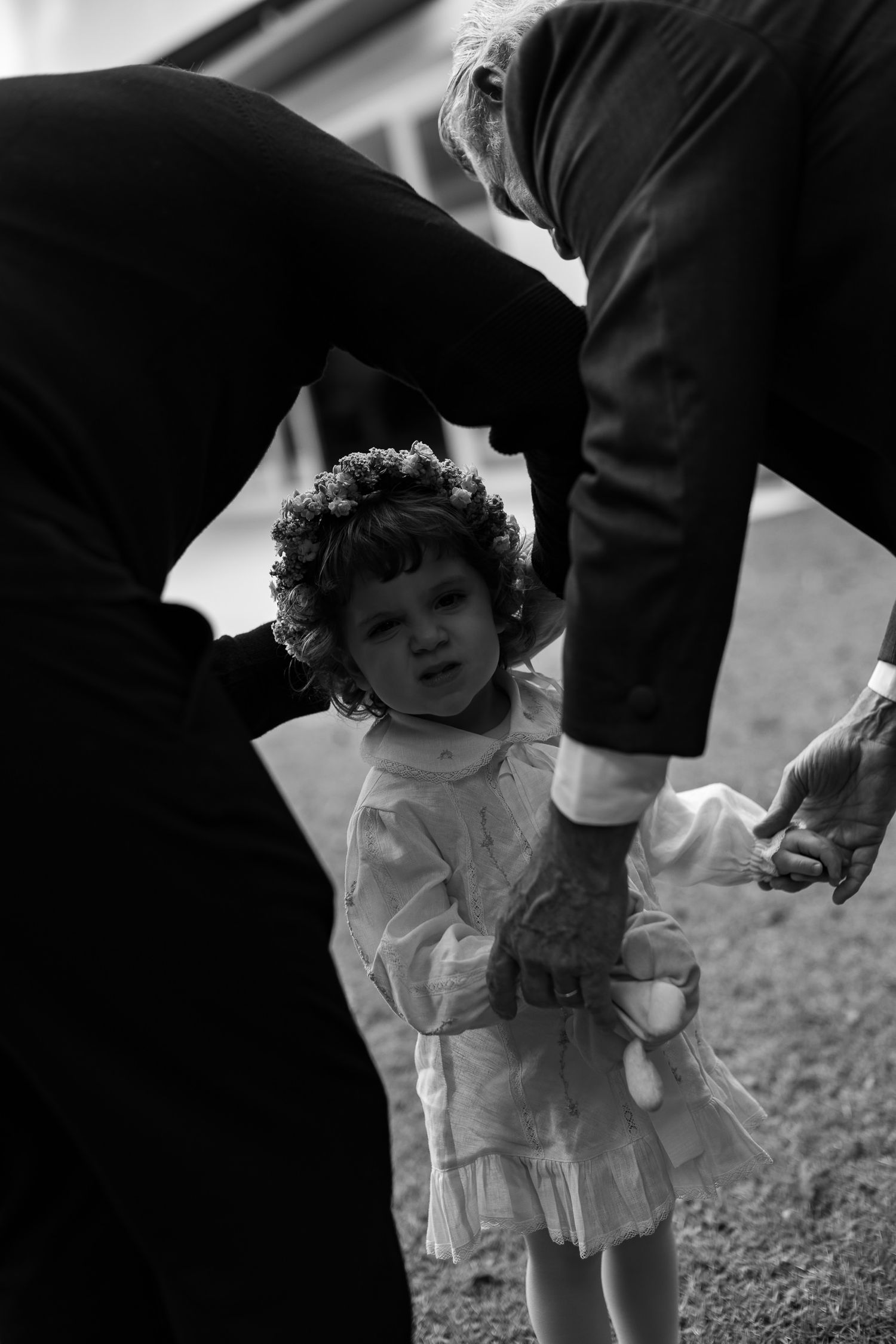 A black and white image shows a small child in a white dress being supported by someone's hands.