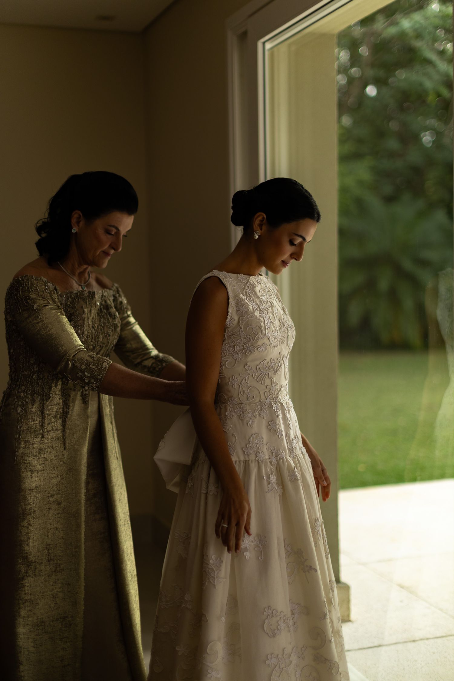 A woman in a white lace wedding dress gets help with final adjustments from someone wearing a gold dress.