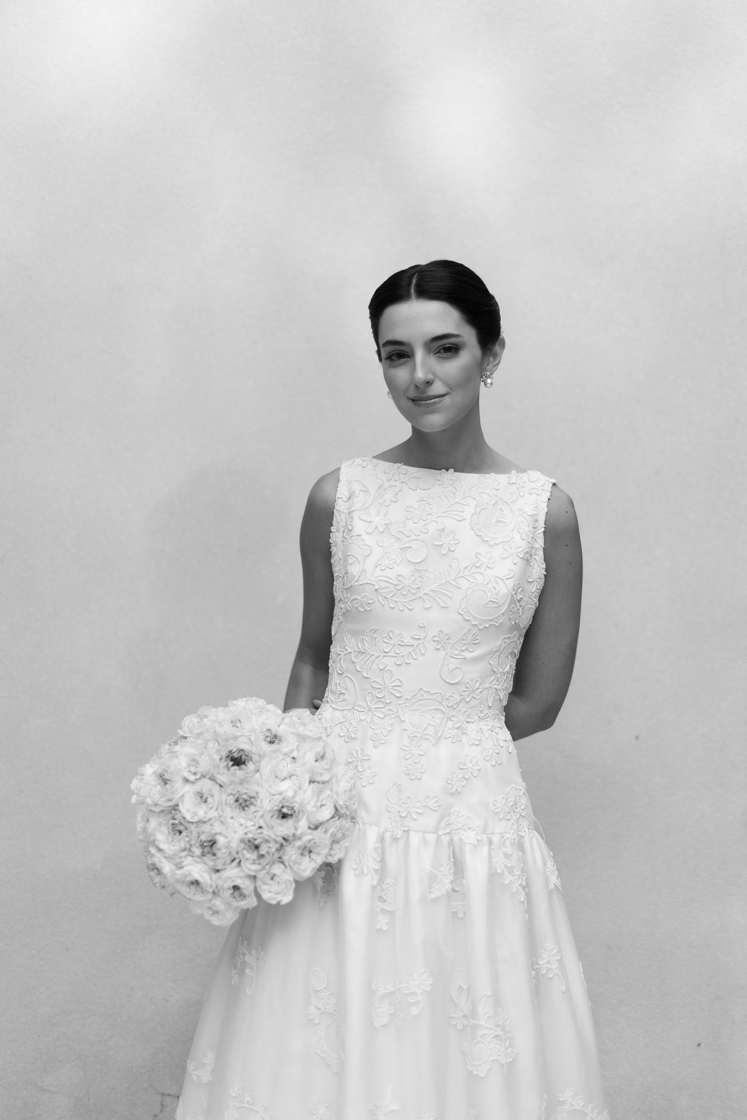 A bride in a sleeveless white lace wedding dress holds a bouquet of white roses against a light background.