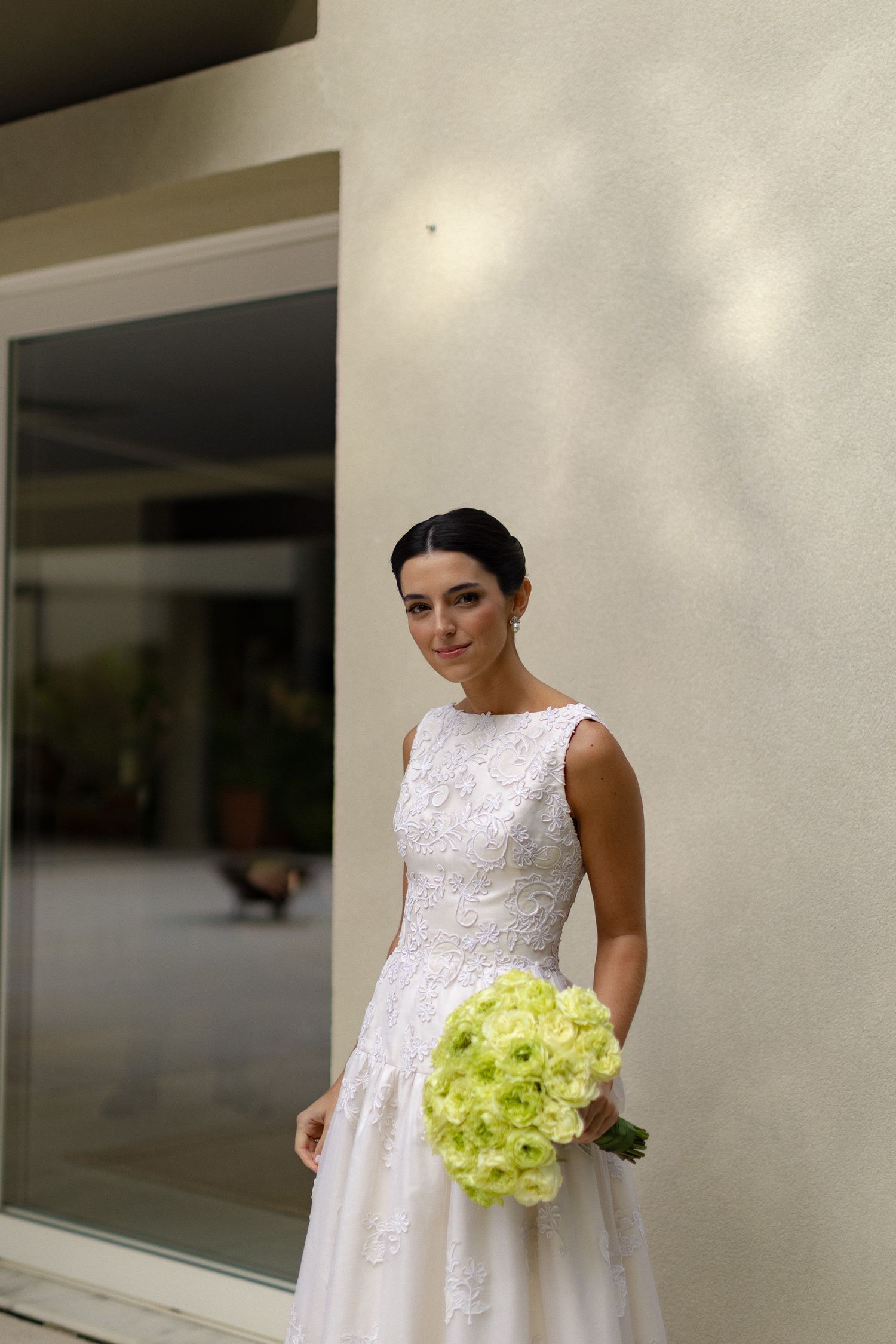 A bride in a classic white sleeveless wedding dress holds a vibrant lime green hydrangea bouquet against a light wall.