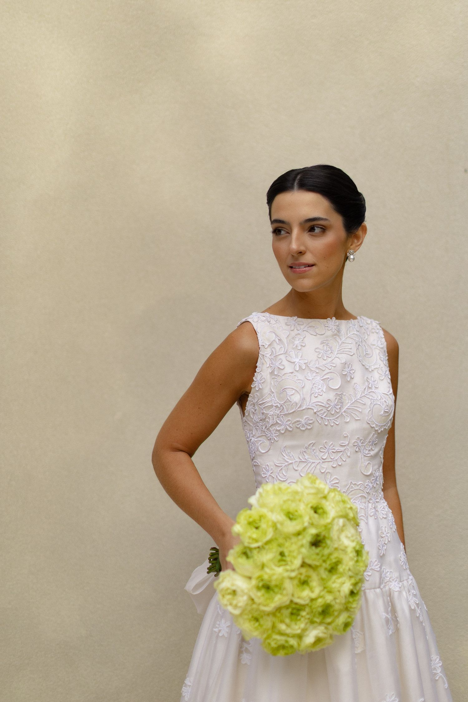 A bride in a sleeveless white lace wedding dress holds a bright lime green hydrangea bouquet against a beige wall.