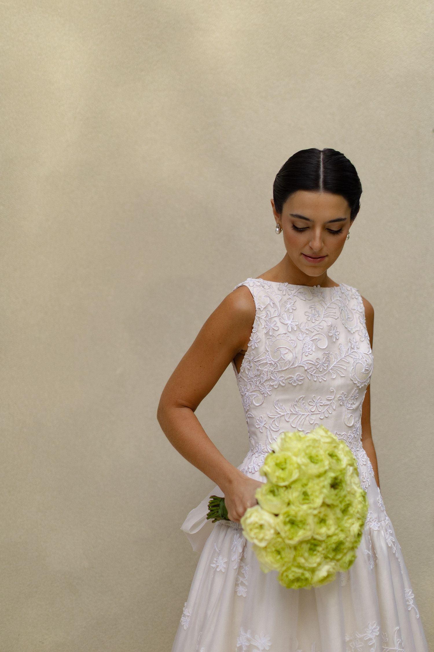 A bride in a sleeveless white lace wedding dress holds a vibrant lime green hydrangea bouquet against a neutral wall.
