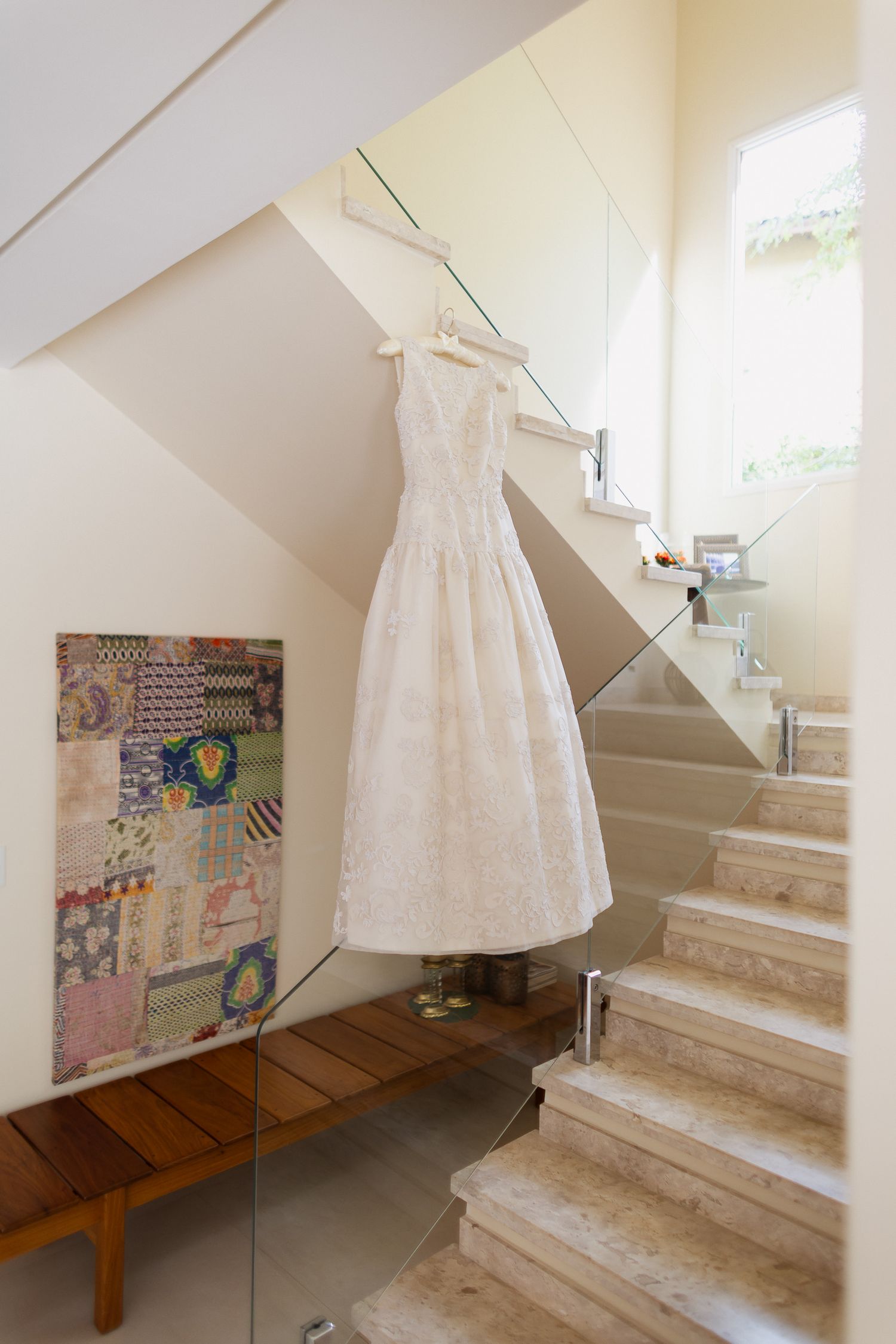 A white lace wedding dress hangs on display in a stairwell next to a patchwork quilt and wooden bench.