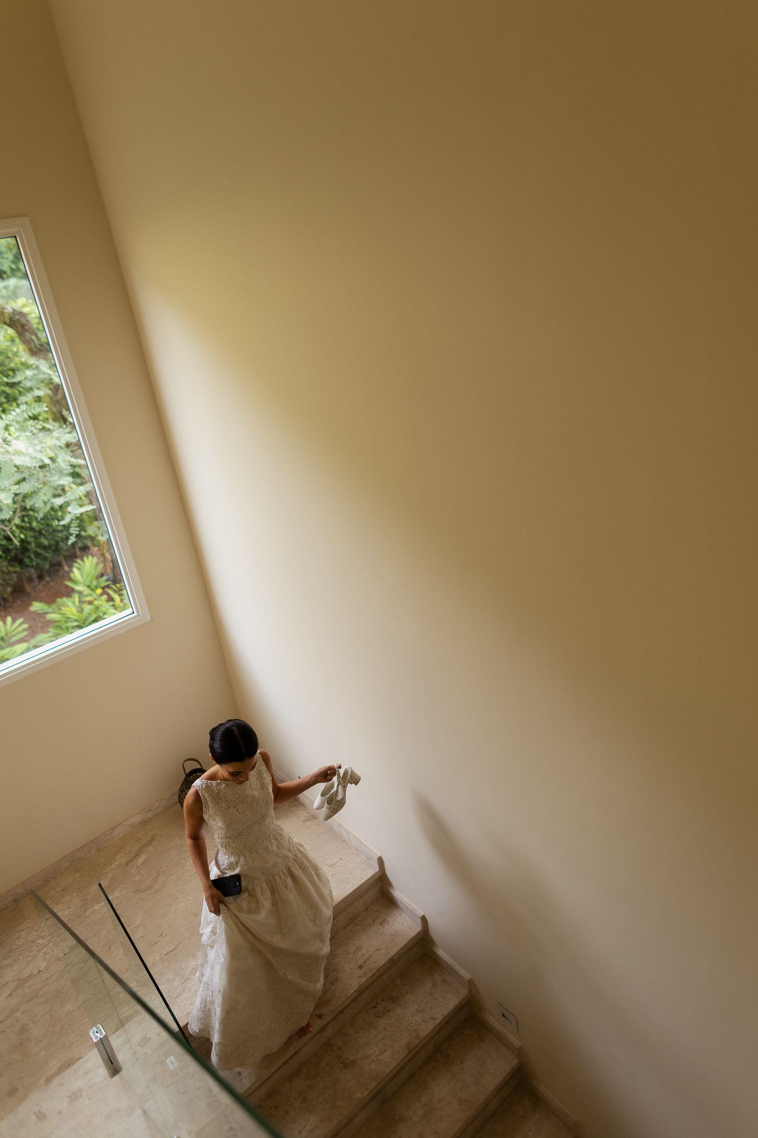 A person in a white dress descends a carpeted staircase in a minimalist interior with a window letting in natural light.