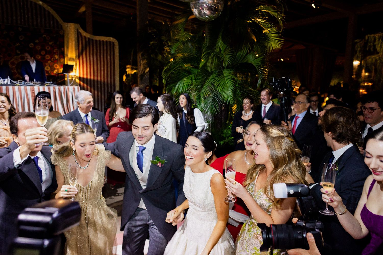 Joyful wedding party dancing and celebrating at an elegant indoor reception with decorative lighting.