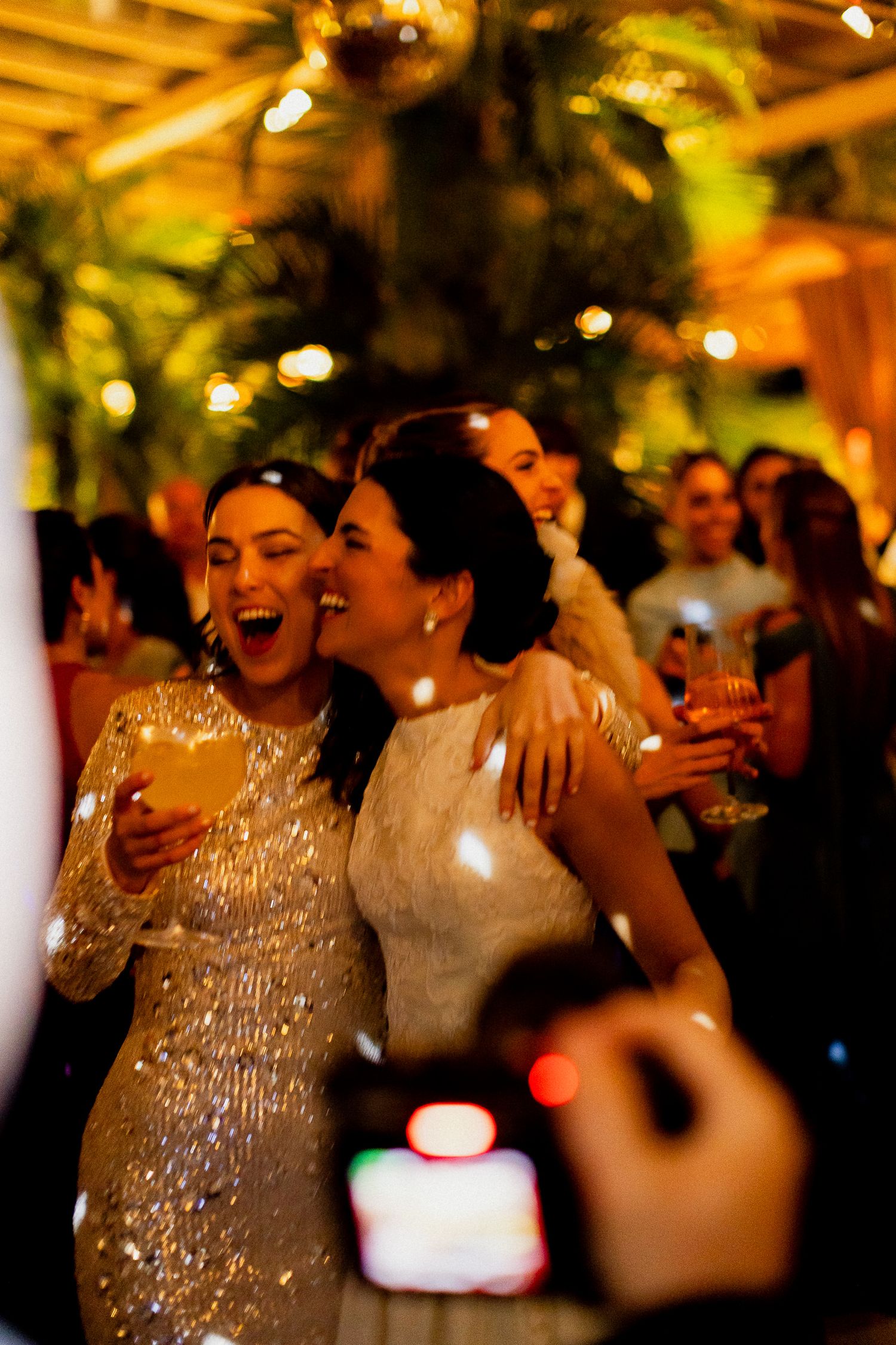 Two people in sparkly formal attire laugh joyfully at an evening celebration with warm lighting and palm decor.