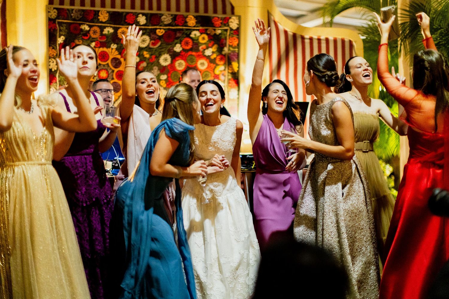 Wedding guests dance and celebrate at a vibrant reception with colorful dresses and festive lighting.