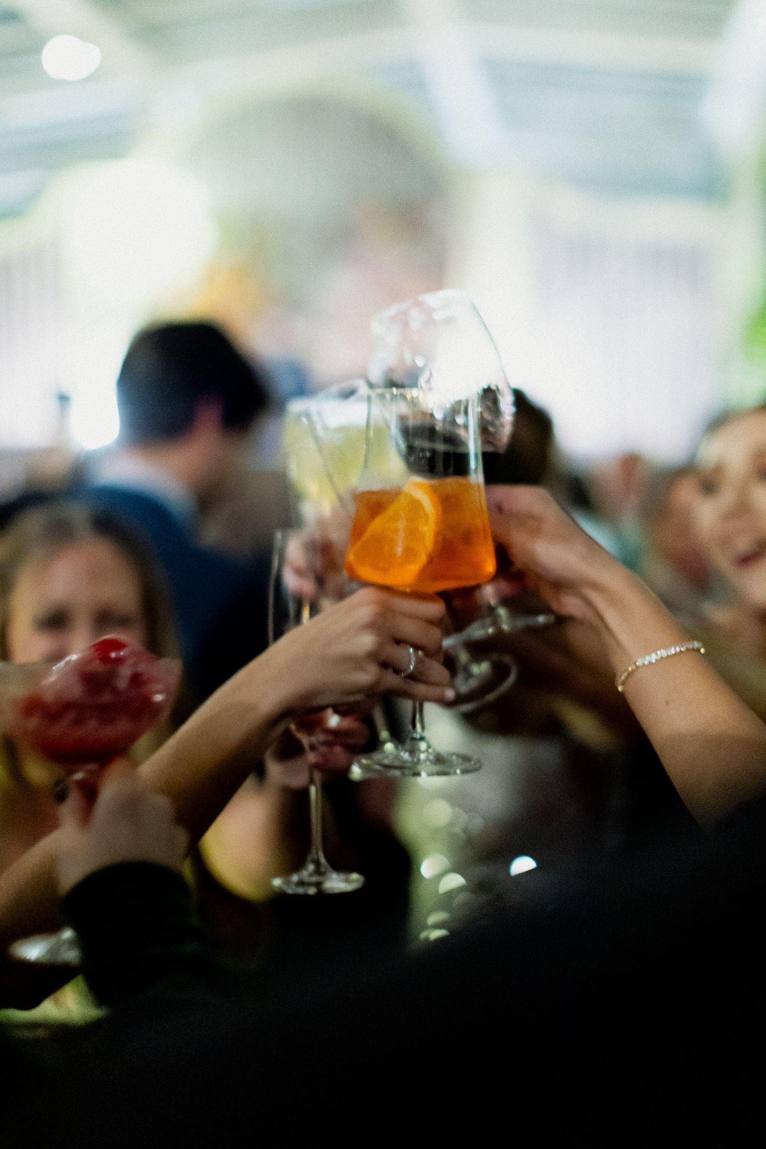 Several hands raise cocktail glasses in a toast during a celebration with an orange-colored drink visible in soft focus.