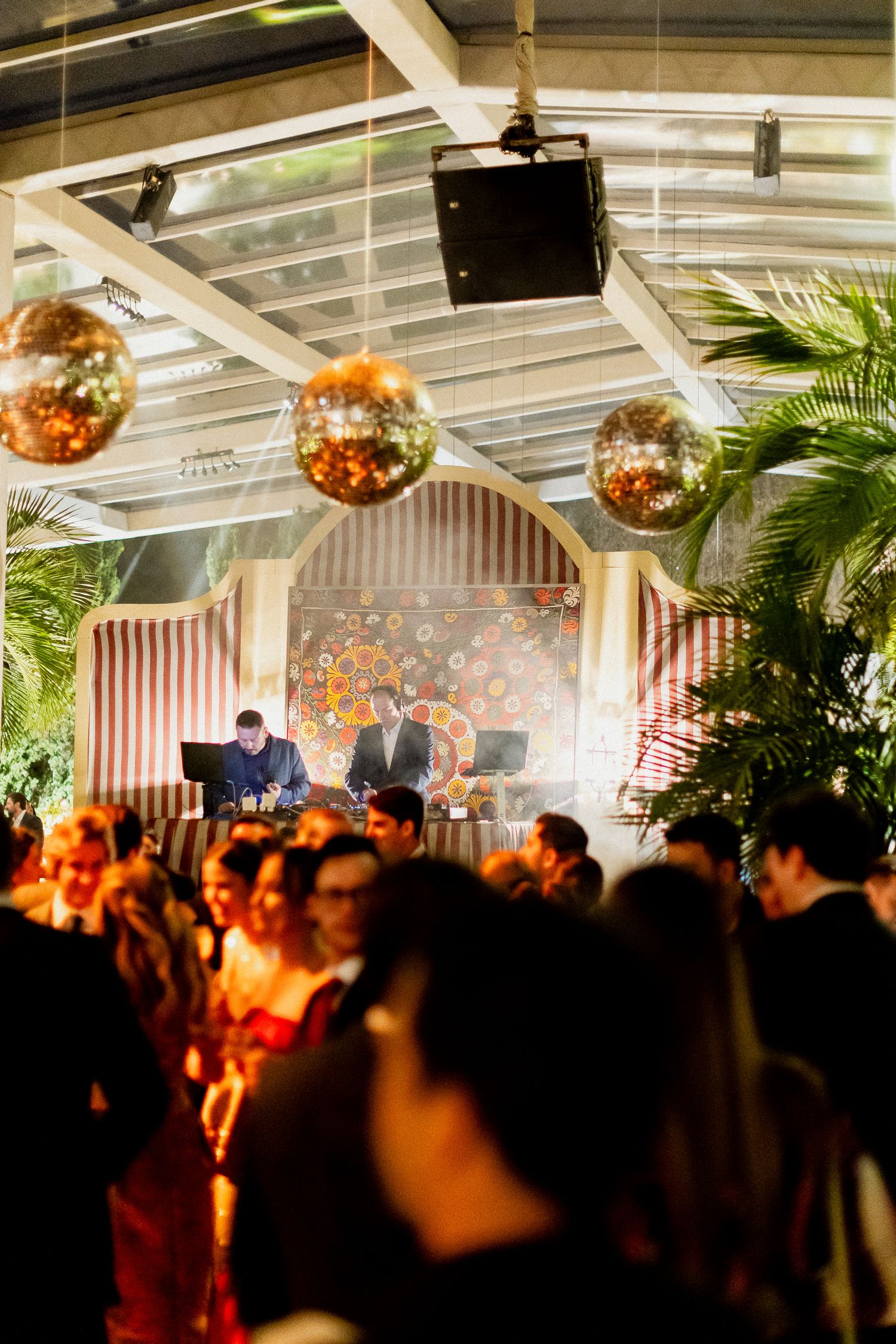 A crowded indoor party venue with disco balls hanging from the ceiling and palm plants decorating the space during an event.