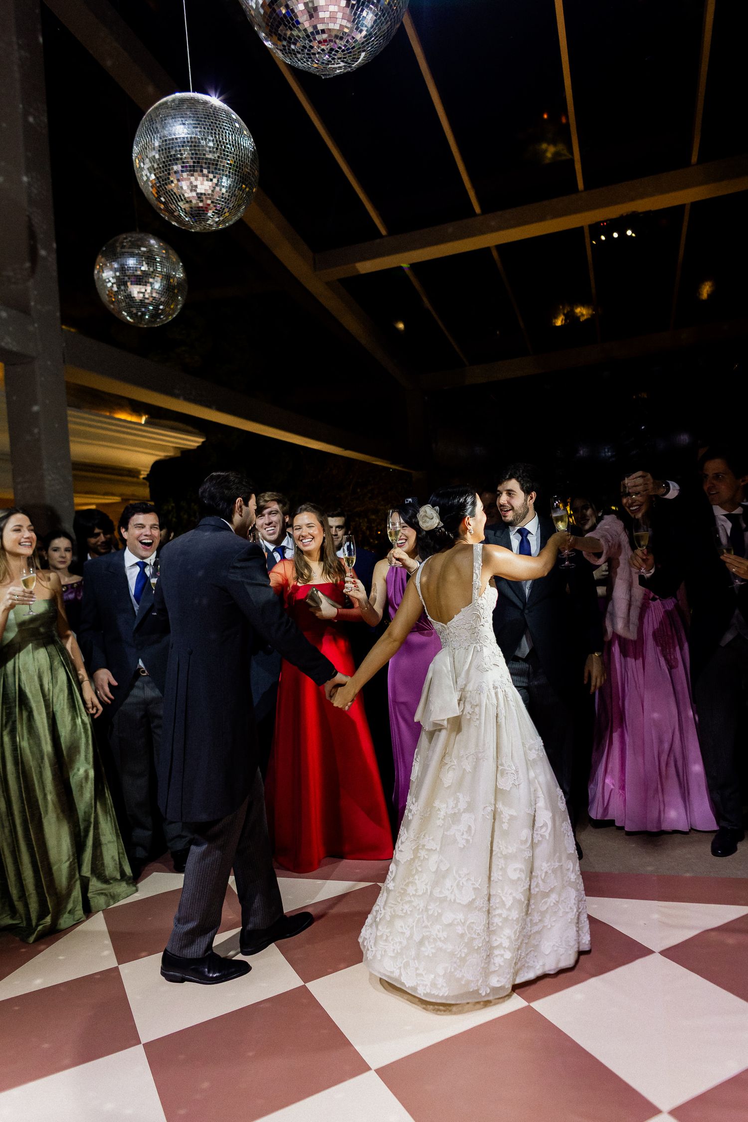 Wedding reception celebration with disco balls overhead and guests dancing on a pink and white checkered dance floor.