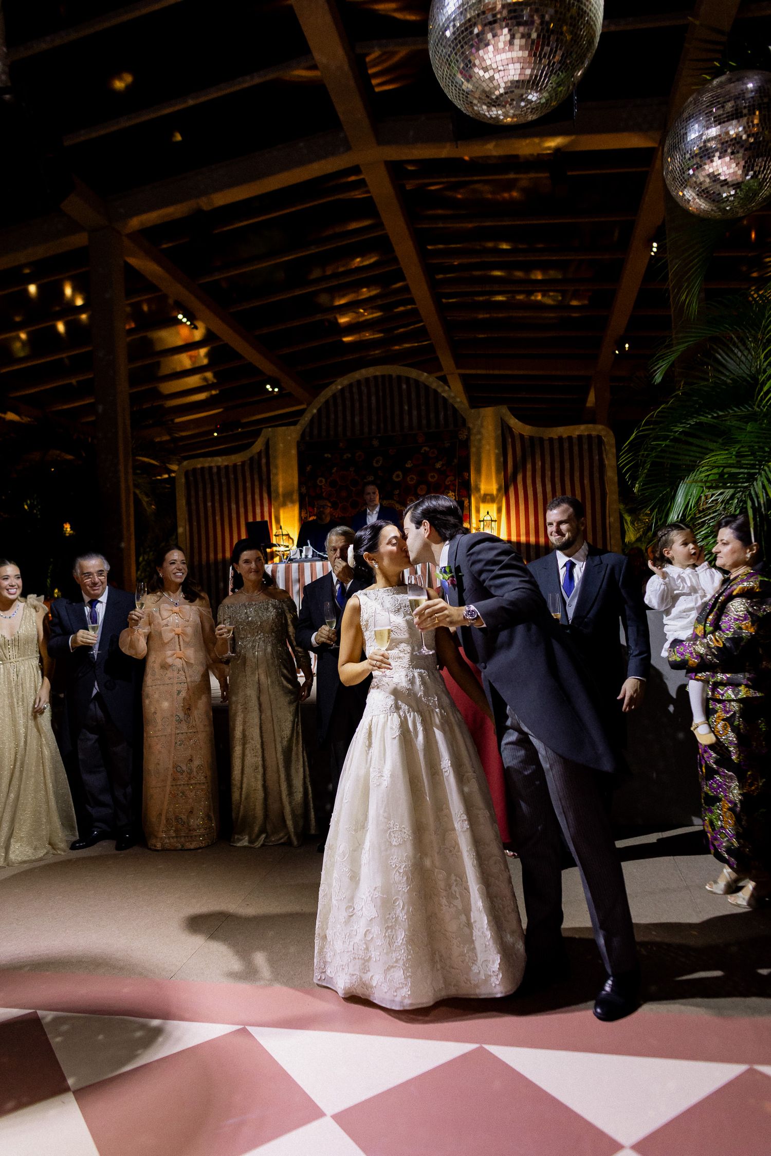 Guests dancing on a pink and white checkered dance floor at an elegant indoor wedding reception with disco balls overhead.