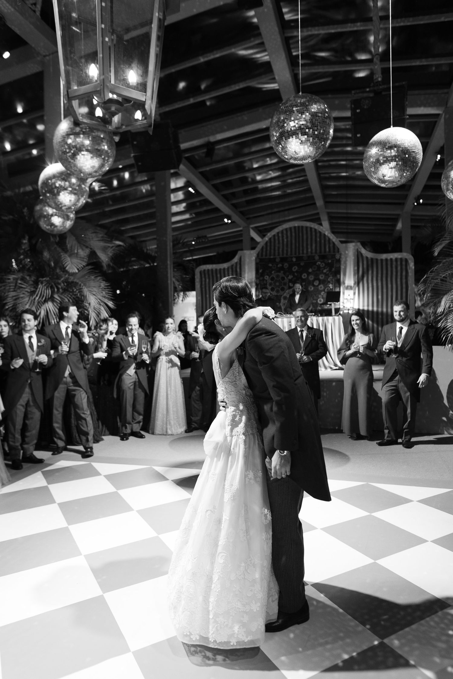 Wedding guests dance together on a black and white checkered dance floor under disco balls at an elegant reception.